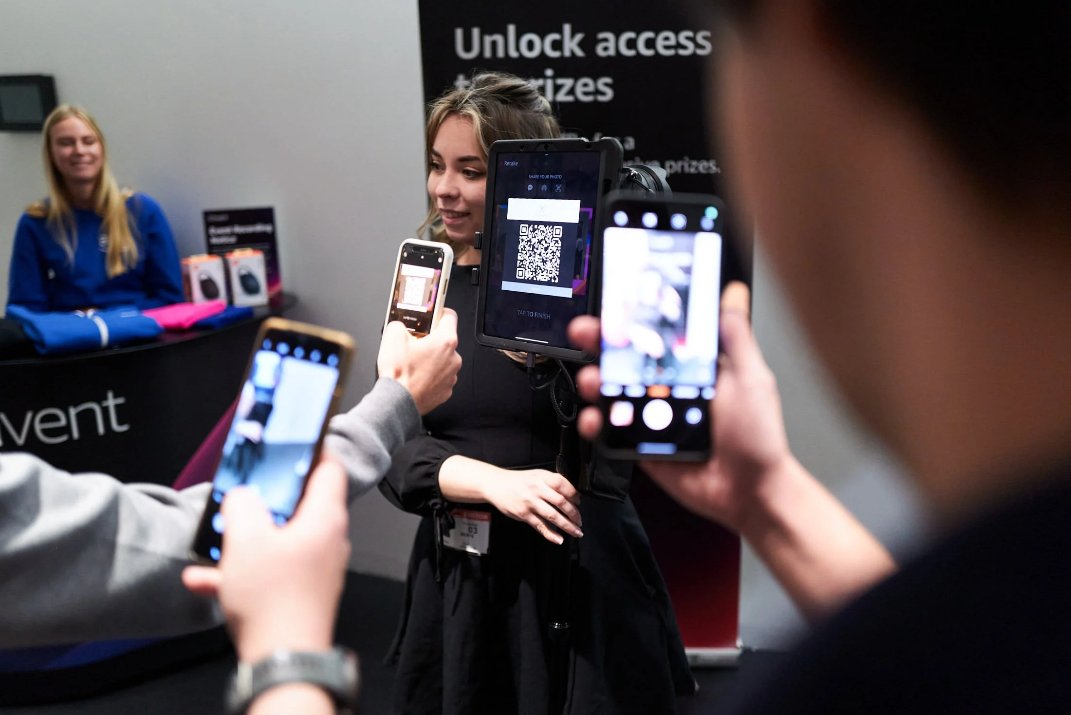 Multiple people taking photos of a woman at a booth with a sign that reads "Unlock access and prizes" in the background.