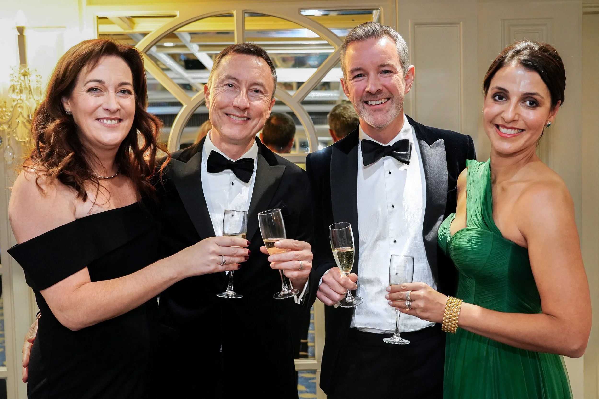 Four adults dressed in formal attire, holding champagne glasses, at a social event or celebration.