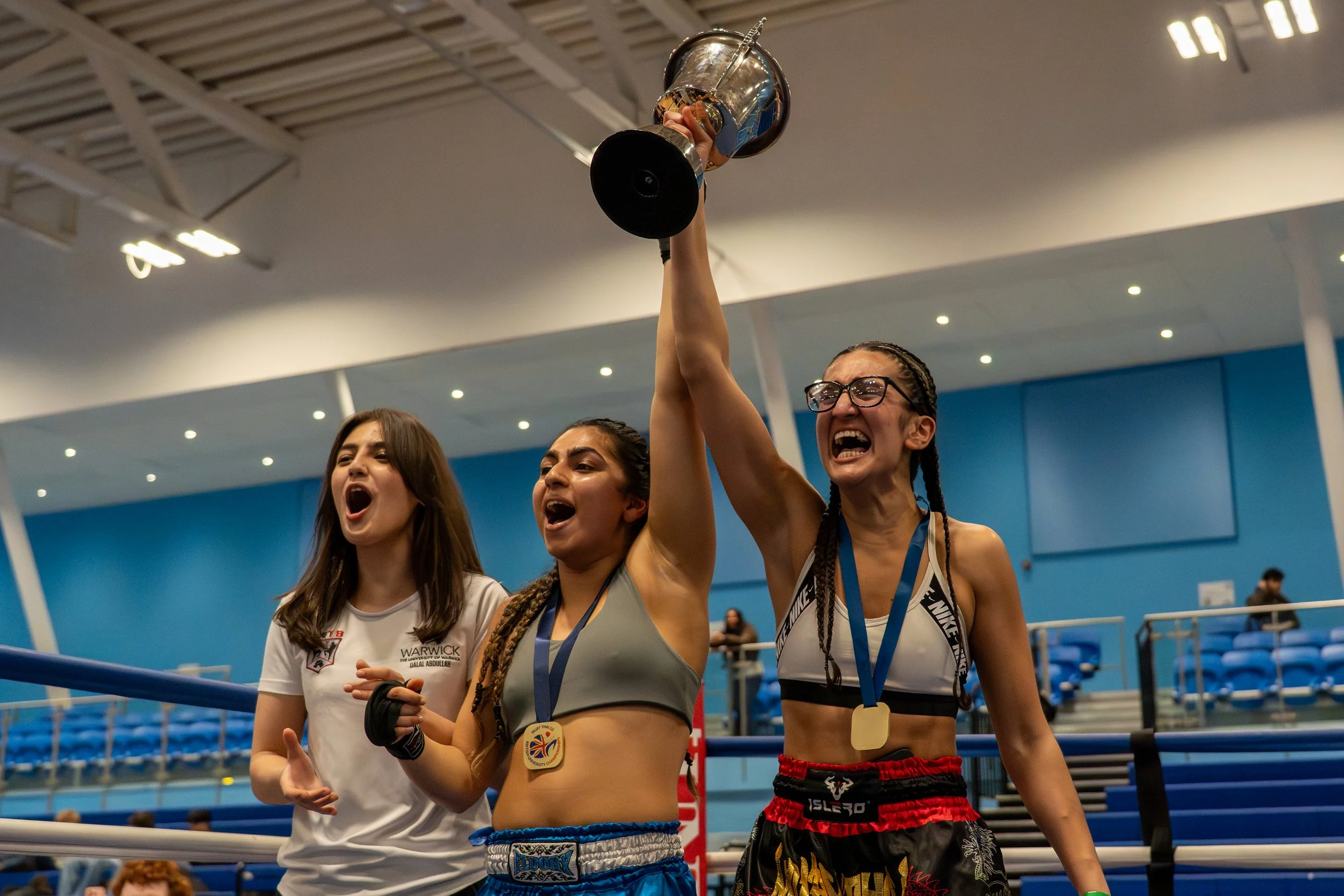 A female boxer celebrating in the boxing ring with a silver trophy, wearing a medals around her neck, and sporting black and red boxing shorts. She is surrounded by other women, with one in a white shirt, inside a sports arena with blue seating.