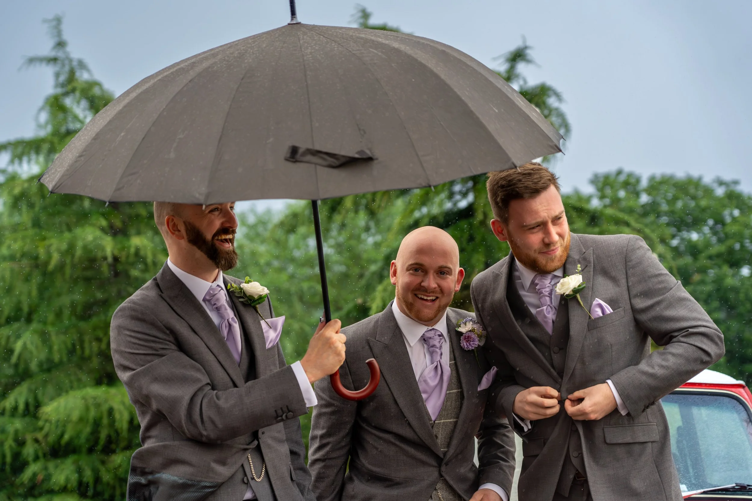 Three men in suits smiling, standing under a large gray umbrella in rainy weather, outdoors with green trees in the background.