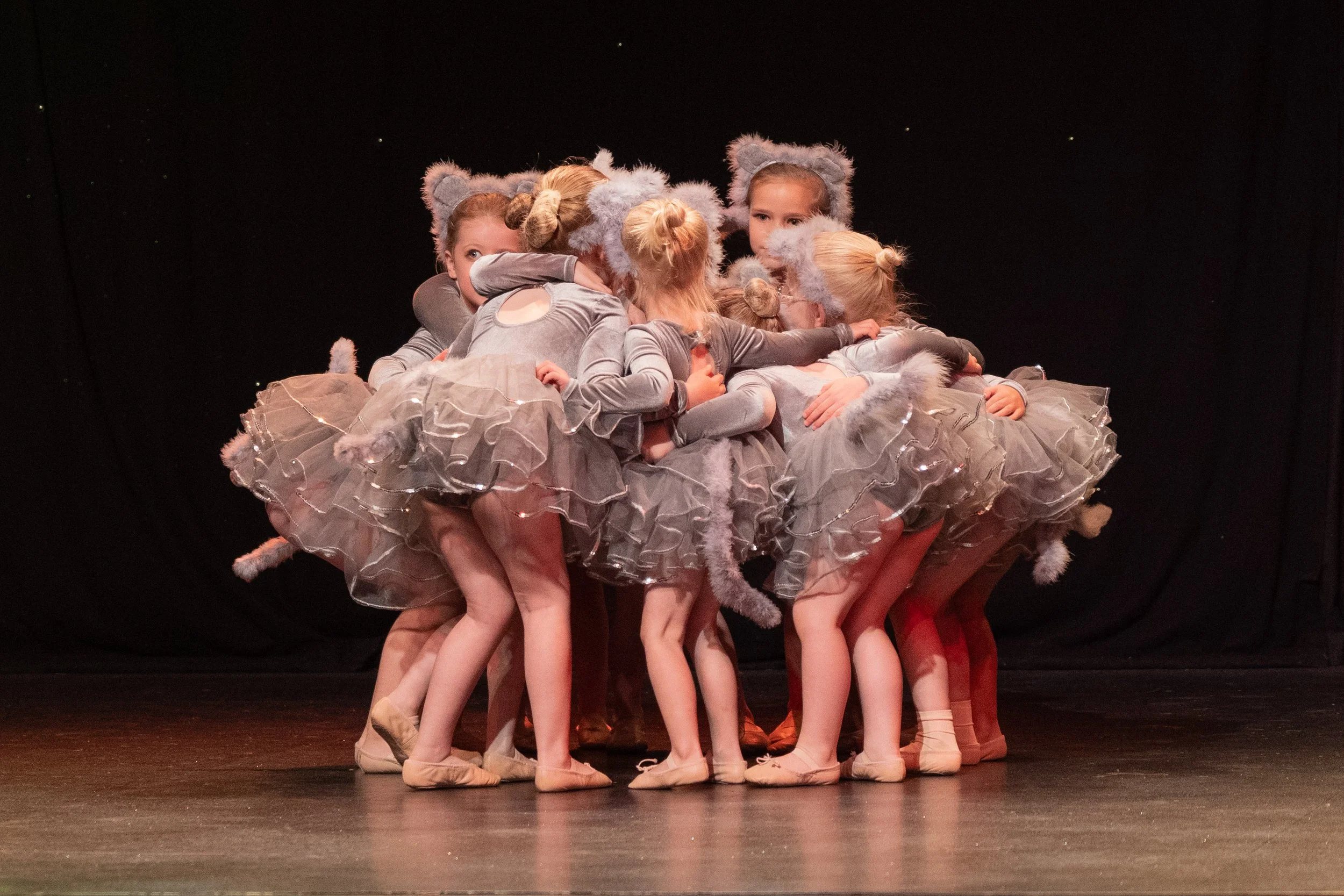 Group of young girls in ballet costumes with gray furry bear ears, huddled together on stage with a black background.