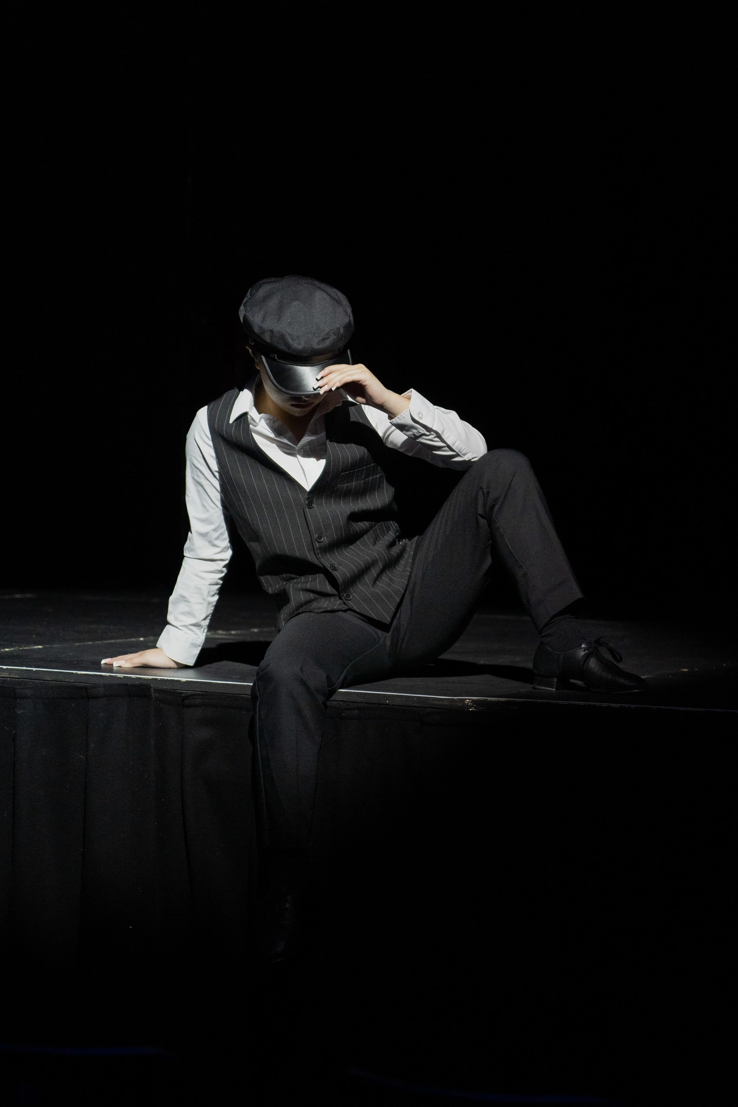 A person sitting on the stage floor with one leg bent and the other extended, wearing a black hat, black vest over a white shirt, and black pants, in a dark setting with a black background.