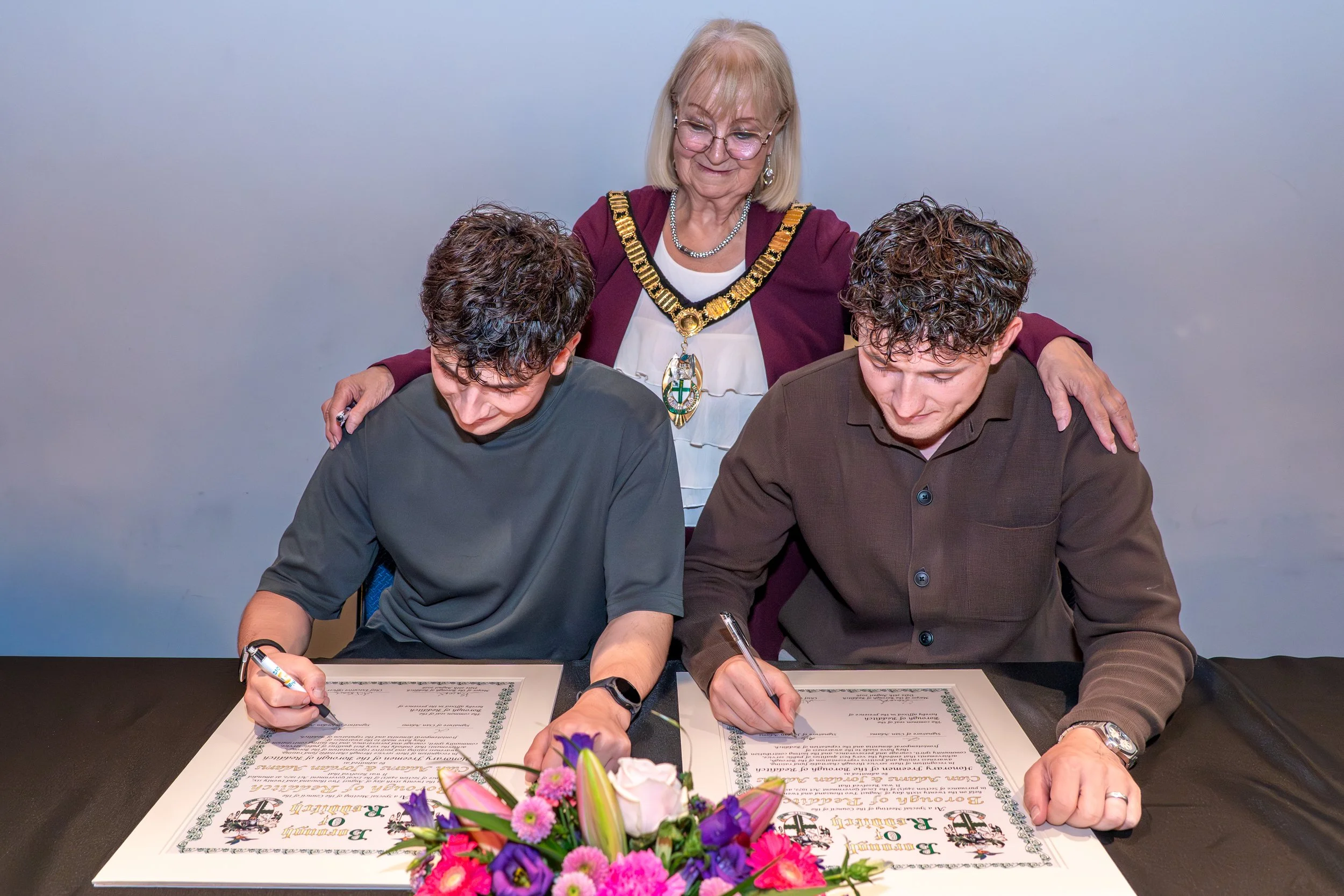 A woman standing behind two men who are signing documents at a table. The woman is wearing glasses, a burgundy jacket, and a ceremonial chain with a medallion. The table has a bouquet of pink, purple, and white flowers. The background is plain and light-colored.
