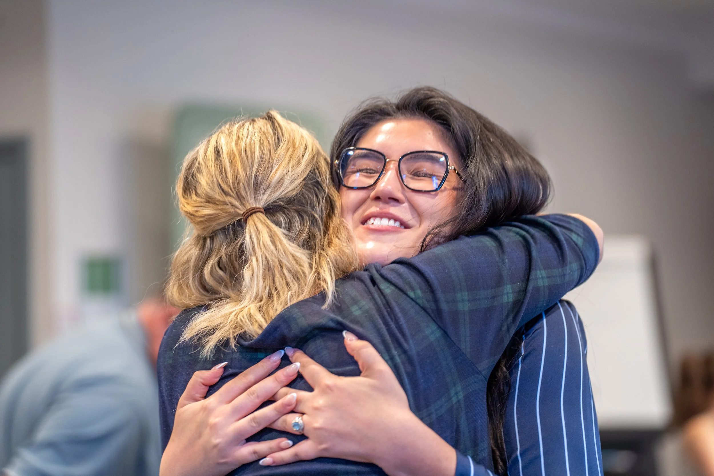 Two women hugging and smiling warmly at each other in an indoor setting.