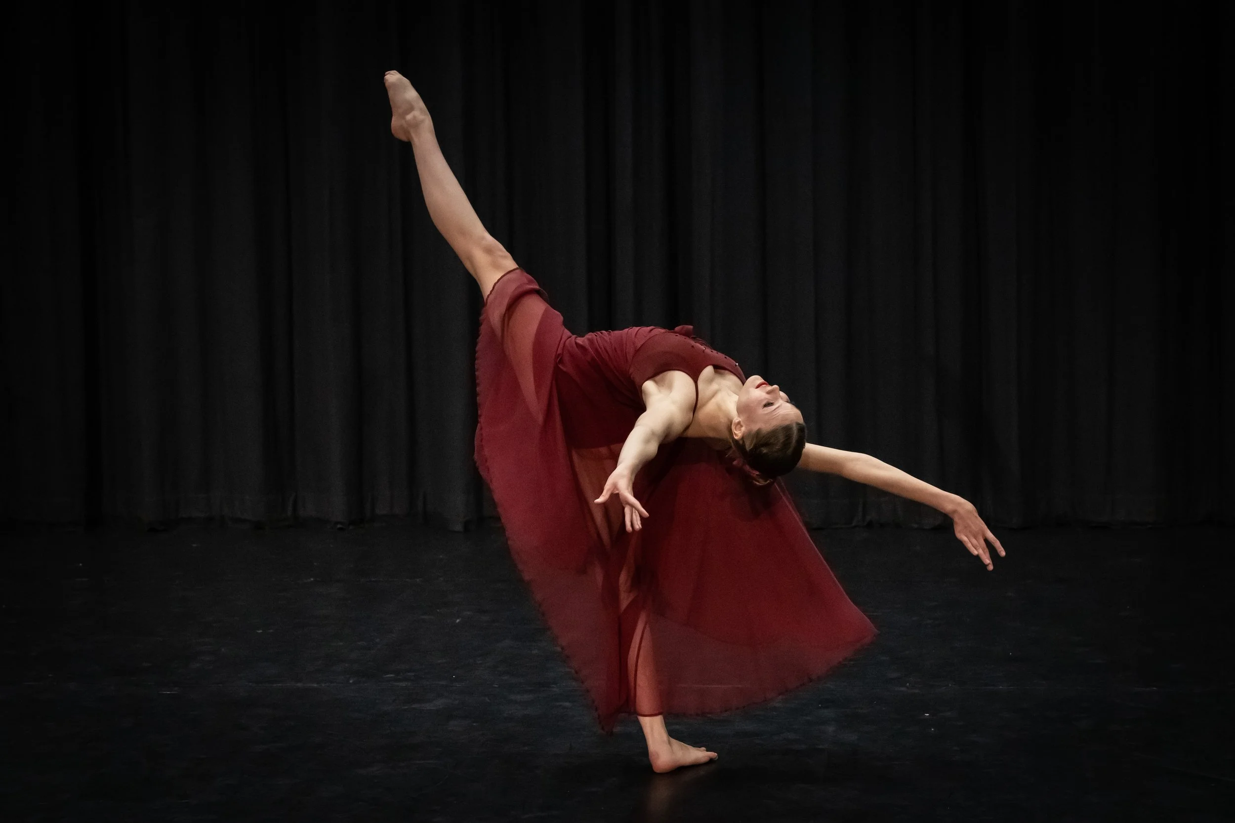 A female dancer in a deep backbend pose on stage, wearing a flowing red dress against a black curtain background.
