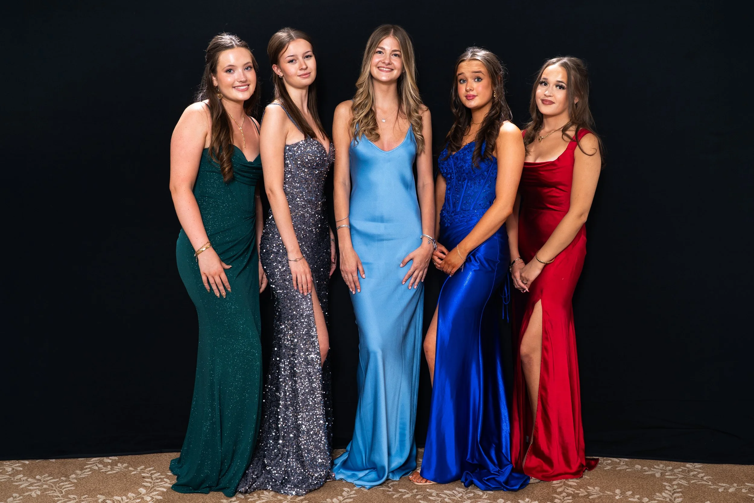 Group of five young women in formal dresses standing together against a black background, smiling.