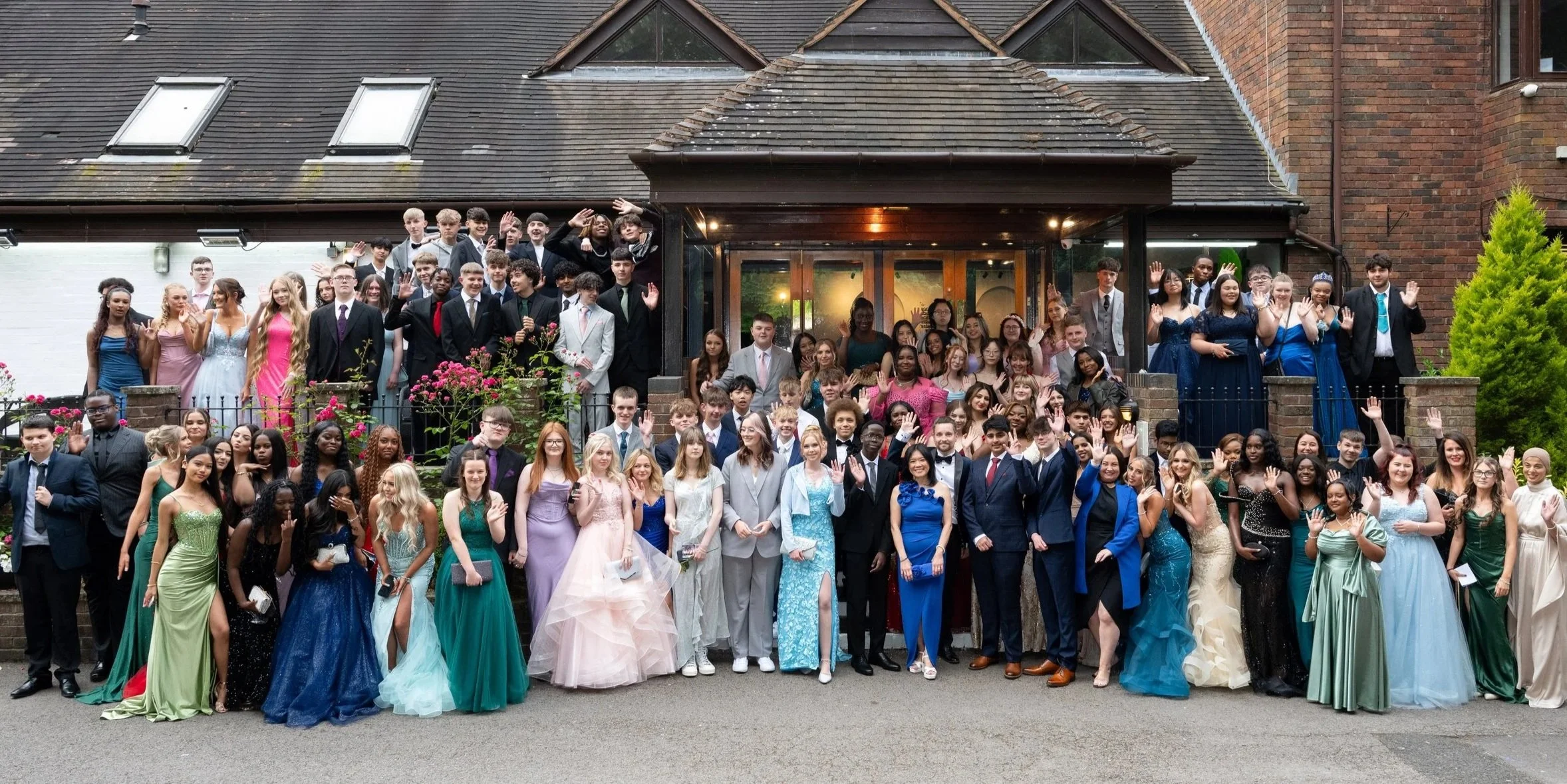 A large group of young people dressed in formal attire, gathered outside a house, waving at the camera. Many are wearing evening dresses and suits, suggesting a celebration or special event.