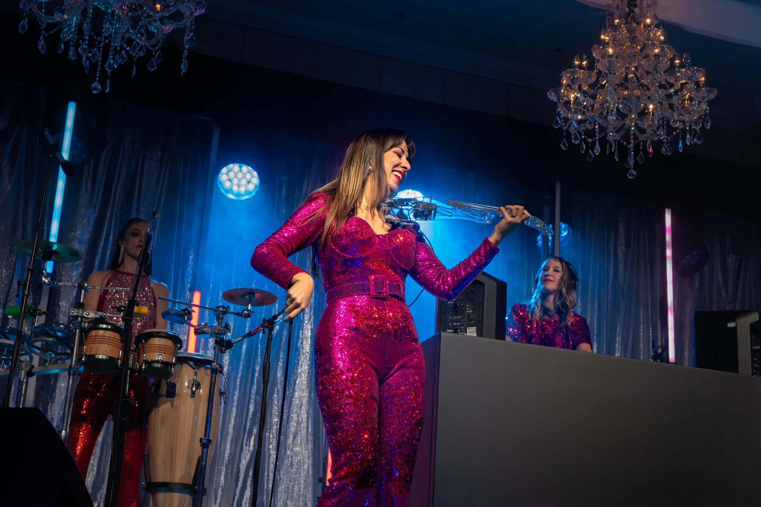 A woman in a shiny pink sequined dress playing a violin on stage with two other women in matching dresses and a percussion instrument in the background, under chandeliers with colorful stage lighting.