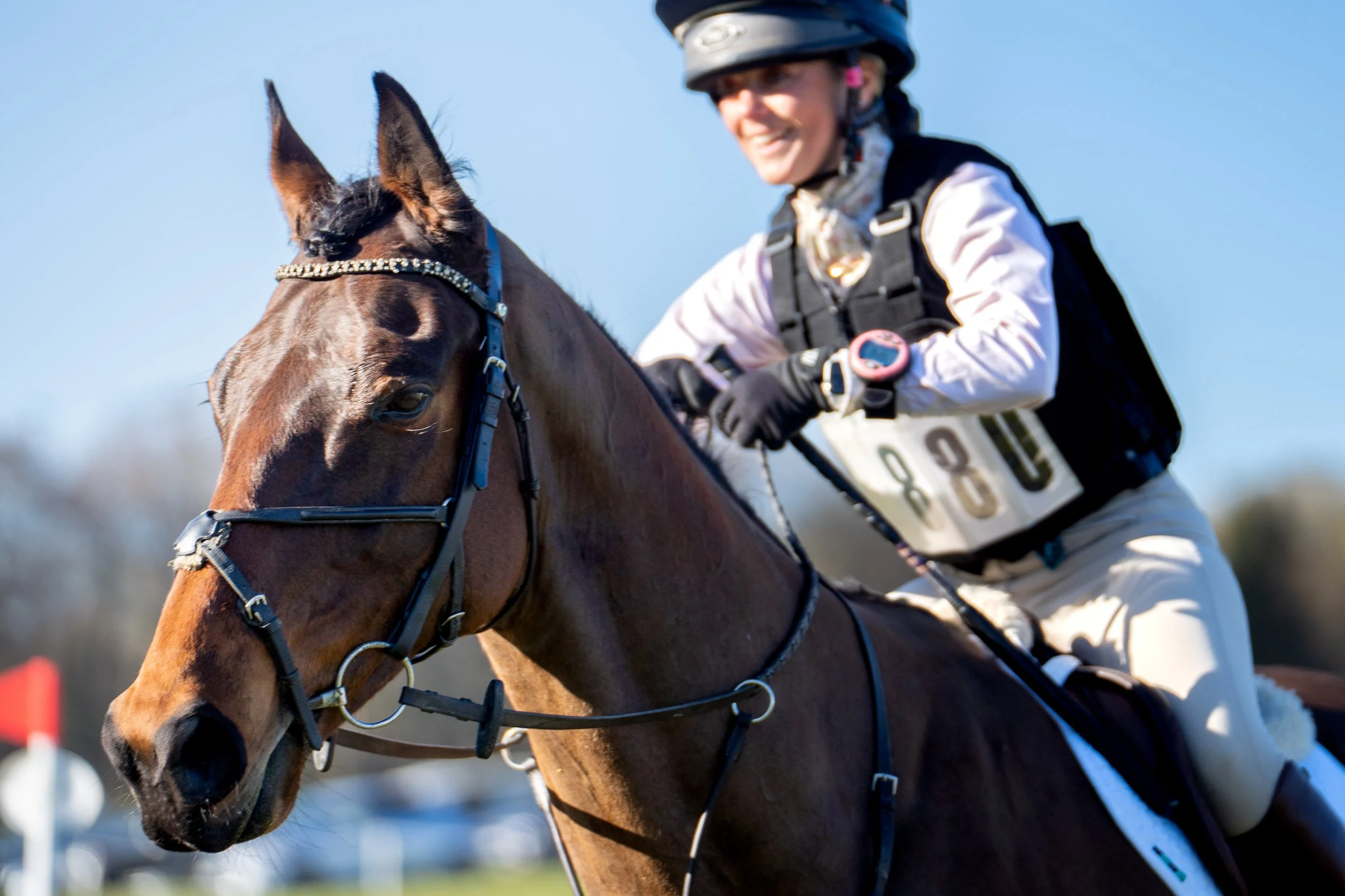 A female jockey riding a brown horse during a horse race, with the jockey smiling and wearing a helmet and riding gear.