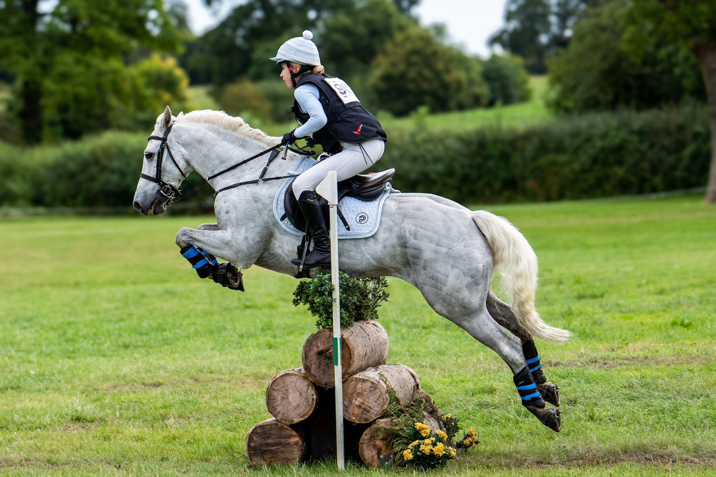 A rider and a white horse jumping over an obstacle made of logs and decorated with yellow flowers during an equestrian event in a grassy field with trees in the background.