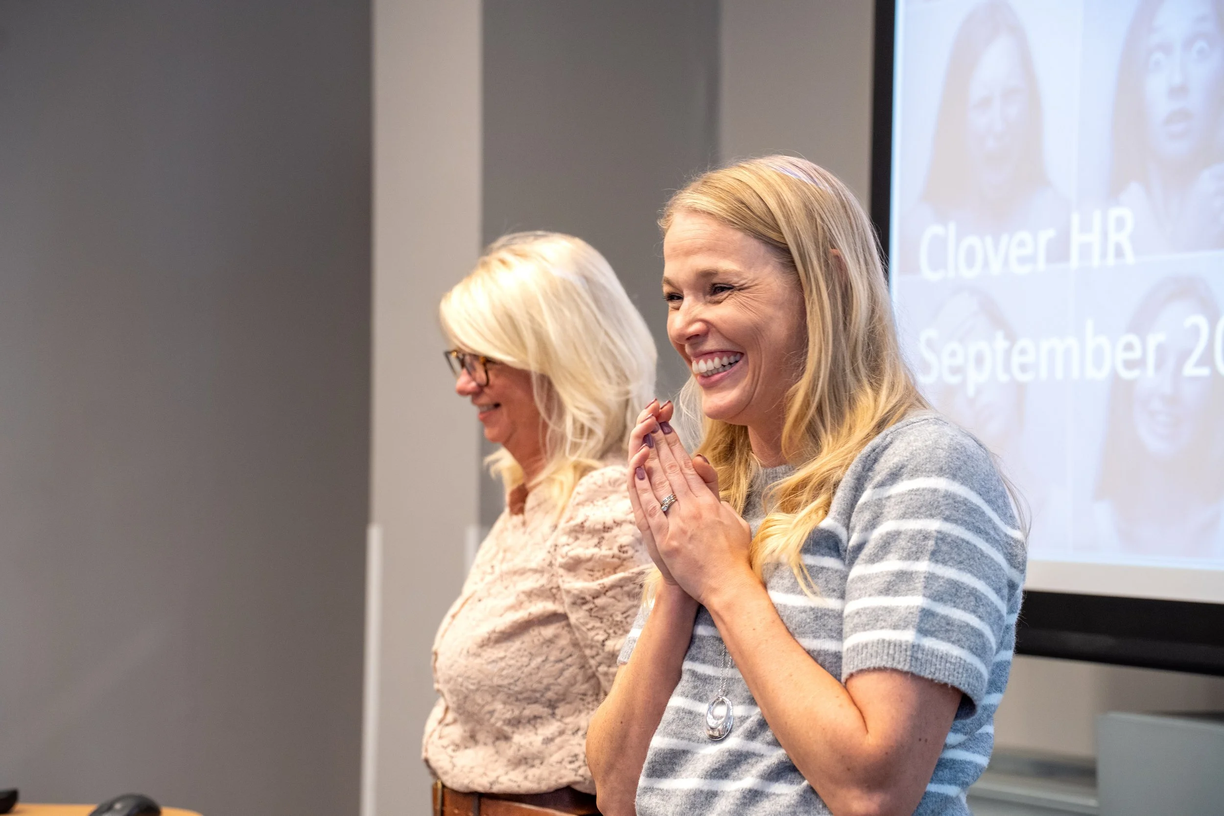 Two women are present in a room, one with blonde hair and glasses, and the other with long blonde hair. They are smiling and appear to be praying or meditating, with their hands clasped together. A presentation slide is visible on the screen behind them, showing the words "Clover HR" and a date in September.
