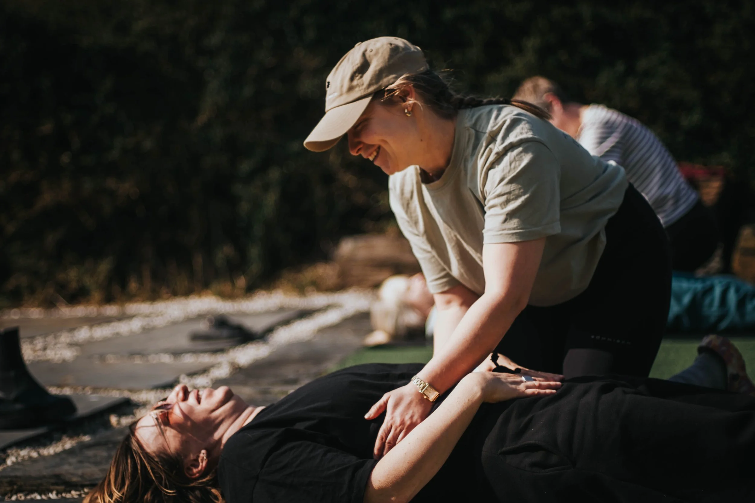 A woman in black lying on her back with eyes closed, receiving CPR from another woman. The woman providing CPR is smiling, wearing a beige cap and a light-colored shirt. They are outdoors with blurred trees in the background.
