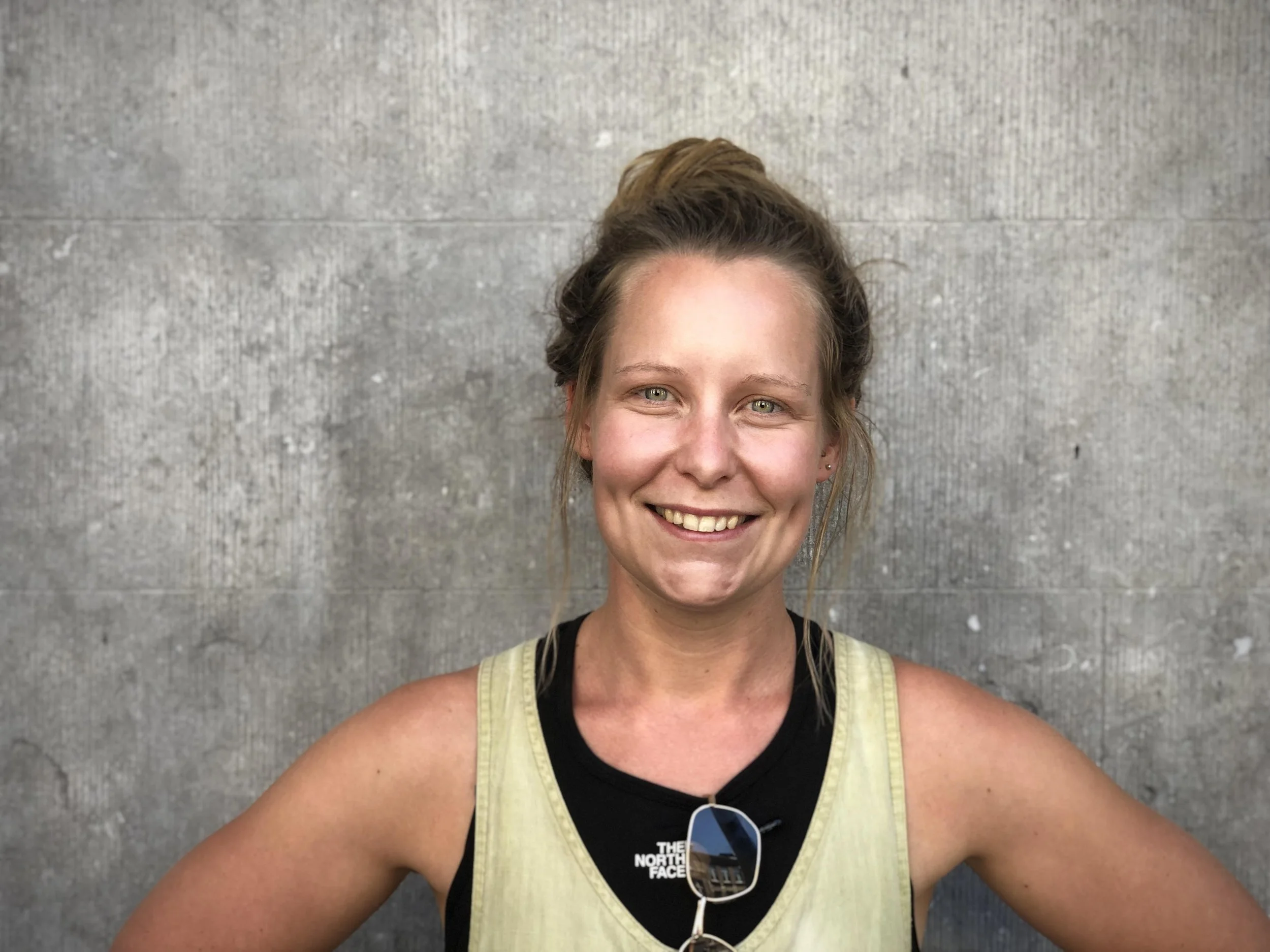 A smiling young woman with light brown hair tied in a bun, wearing a yellow tank top and sunglasses hanging on her chest, taking a selfie against a concrete wall.