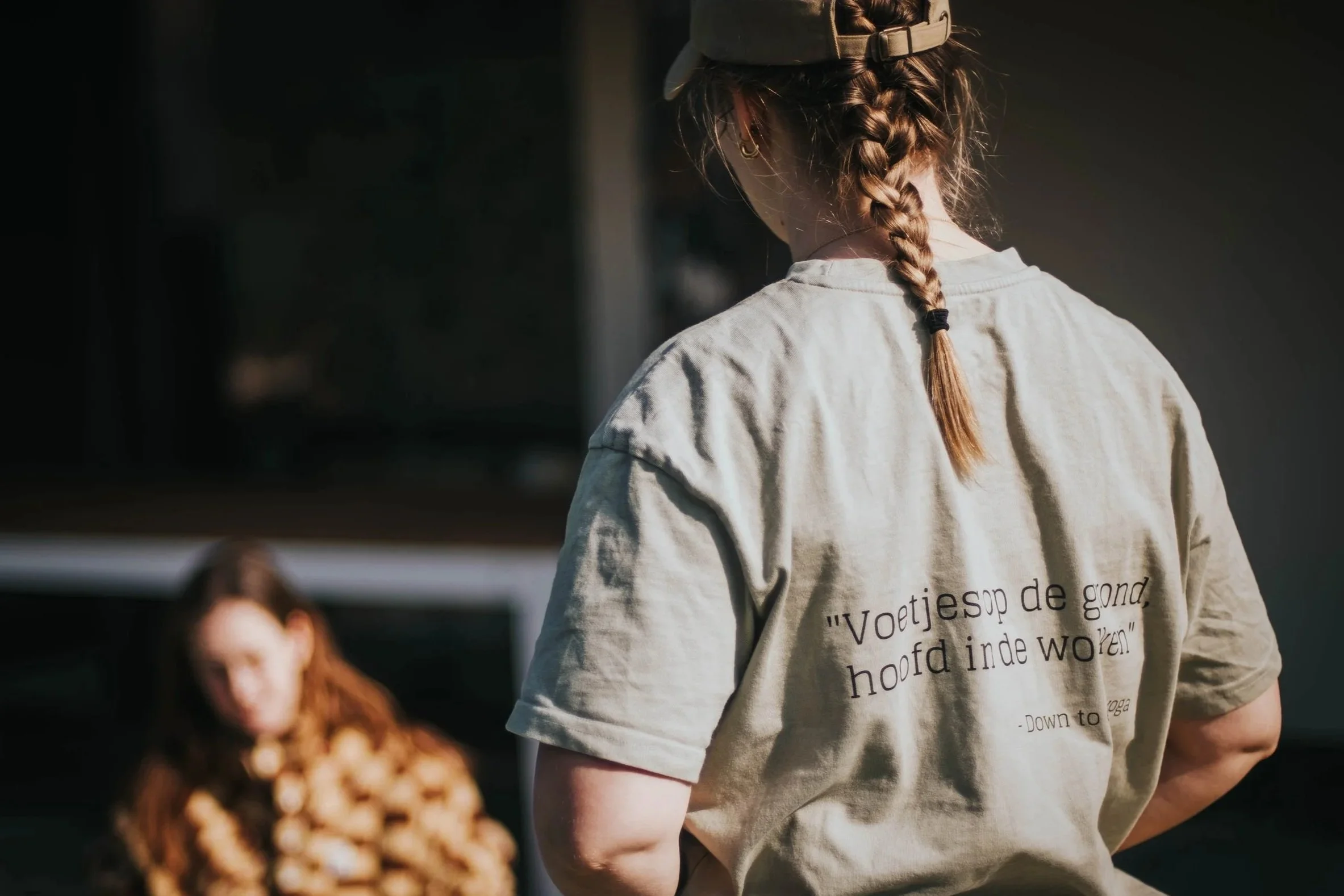 A person with braided hair wearing a beige baseball cap and a beige T-shirt with black text on the back, standing beside a child with long curly red hair.