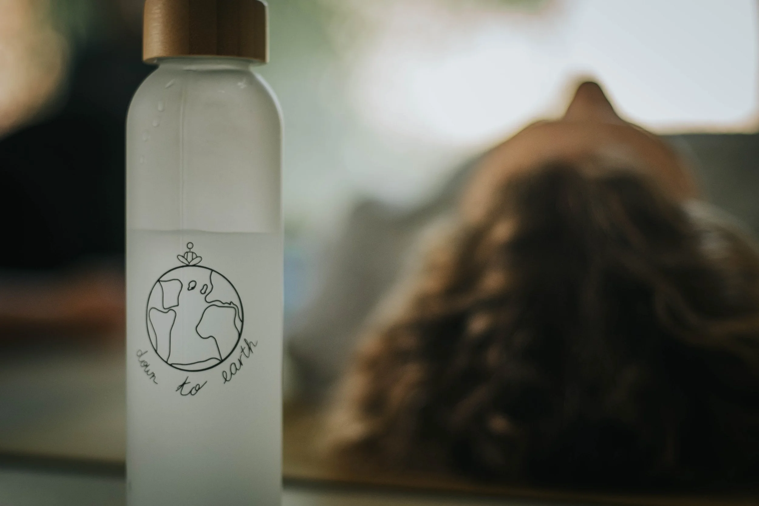 A white bottle with a black illustration of the Earth and the words "to earth" written around it. The bottle is placed on a surface with a blurred background and a person with long dark hair visible in the foreground.