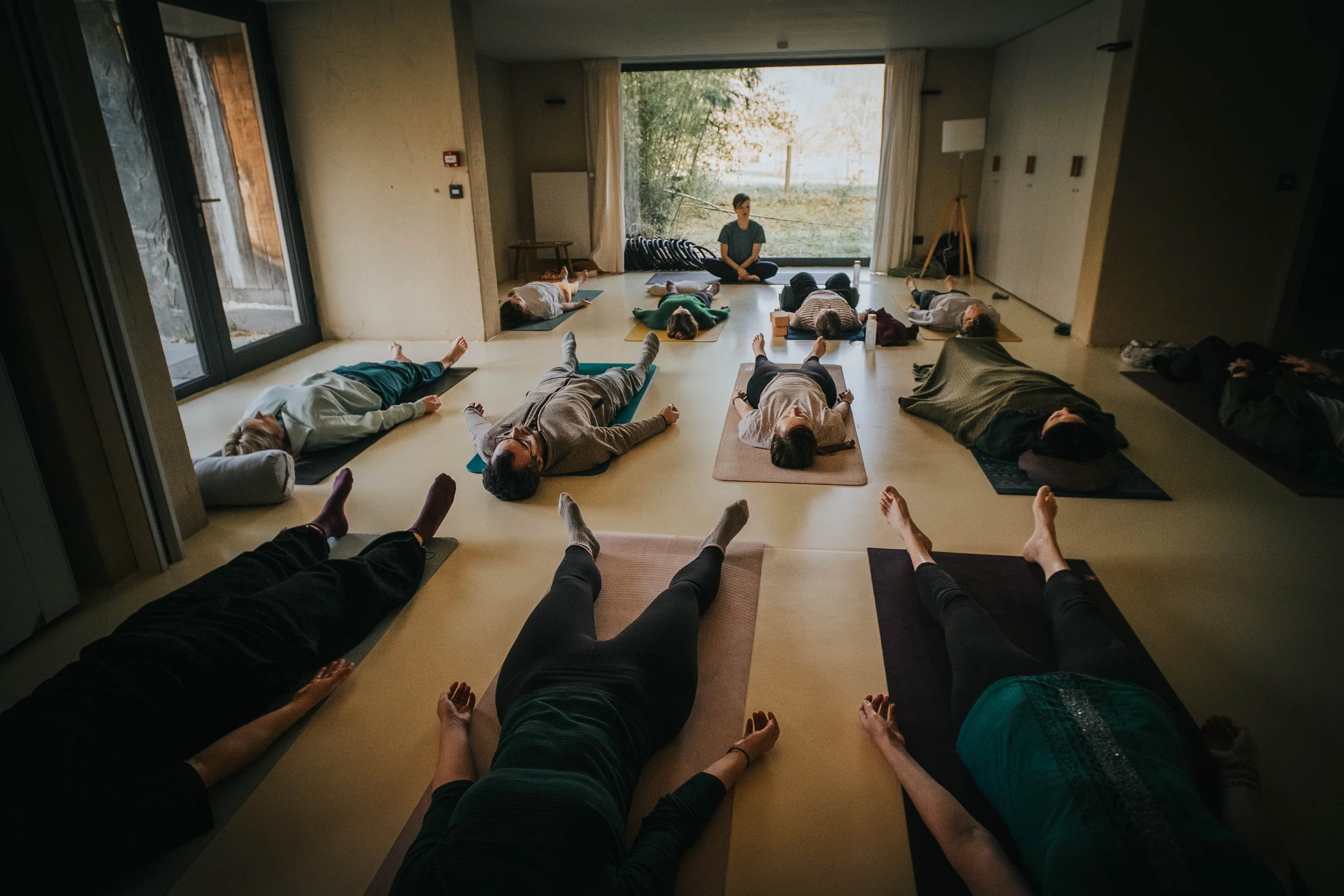 People participating in a yoga class, lying on mats in a spacious room with large window showing outdoor greenery, instructor sitting in front.