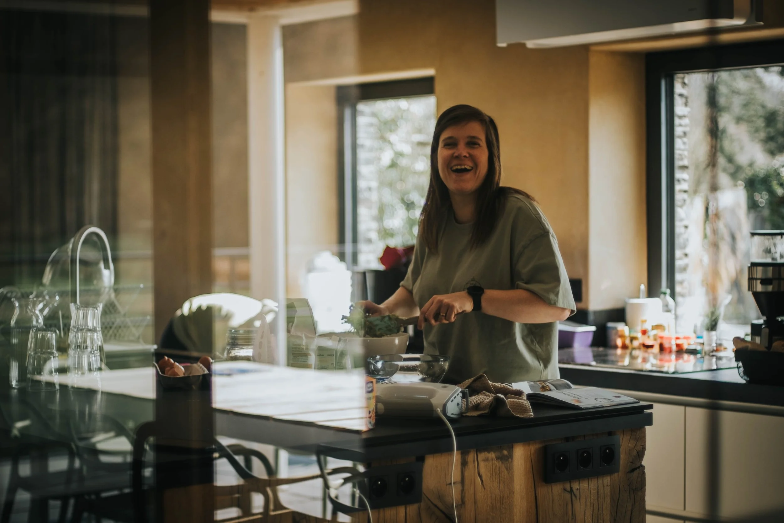 A woman with dark hair smiling and laughing while cooking in a modern kitchen with large windows and natural light. She is wearing a casual t-shirt and standing at the kitchen counter.