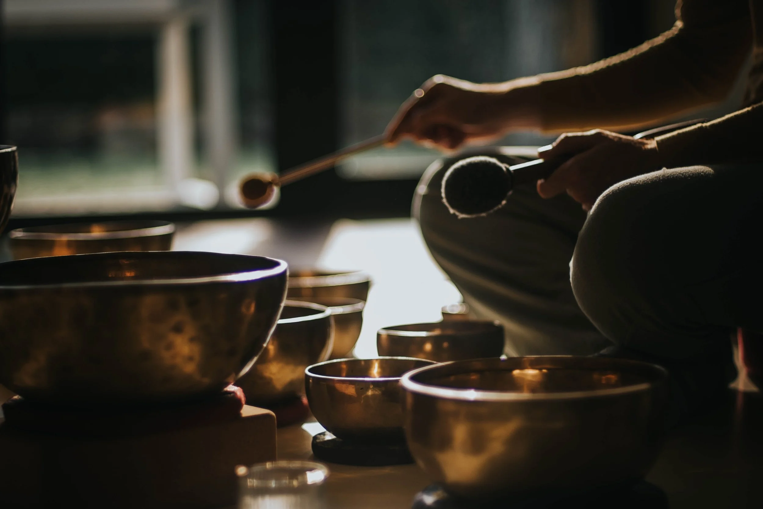 Person pouring a dark liquid from a spoon into a small metallic bowl, with several other metallic bowls in the foreground, in a dimly lit room.