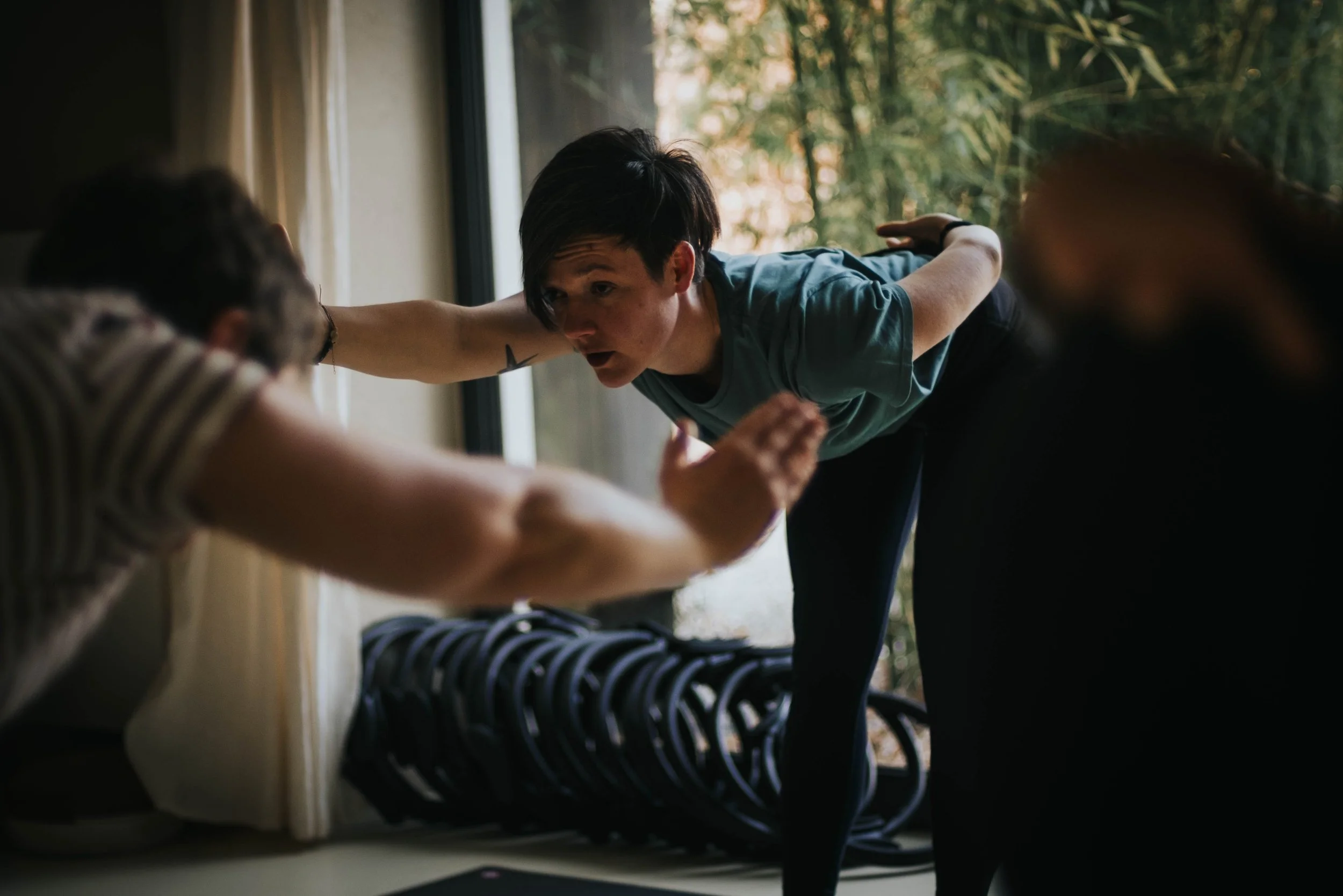 A woman is practicing yoga or stretching near a large window with trees outside, with her arm extended and her gaze focused.