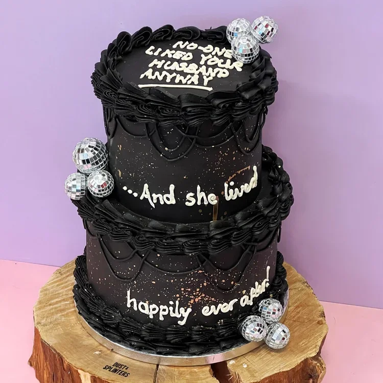 a black two tiered cake with retro icing saying 'no one liked your husband anyway' decorated with disco balls