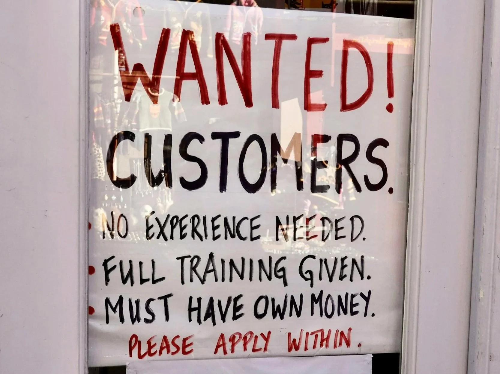 Sign on a door or window that reads 'WANTED! CUSTOMERS. NO EXPERIENCE NEEDED. FULL TRAINING GIVEN. MUST HAVE OWN MONEY. PLEASE APPLY WITHIN.'