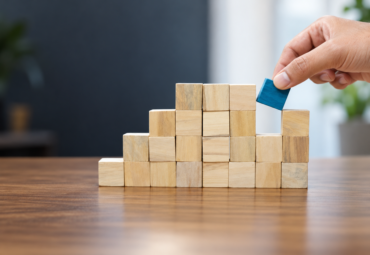 A hand placing a blue wooden block on a pyramid made of natural wood blocks on a wooden table.