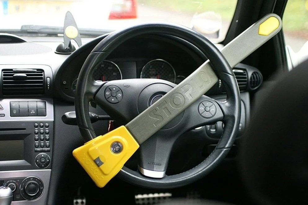 Inside a car, a steering wheel with a large gray and yellow STOP sign attached across it. The dashboard and instrument panel are visible in the background.