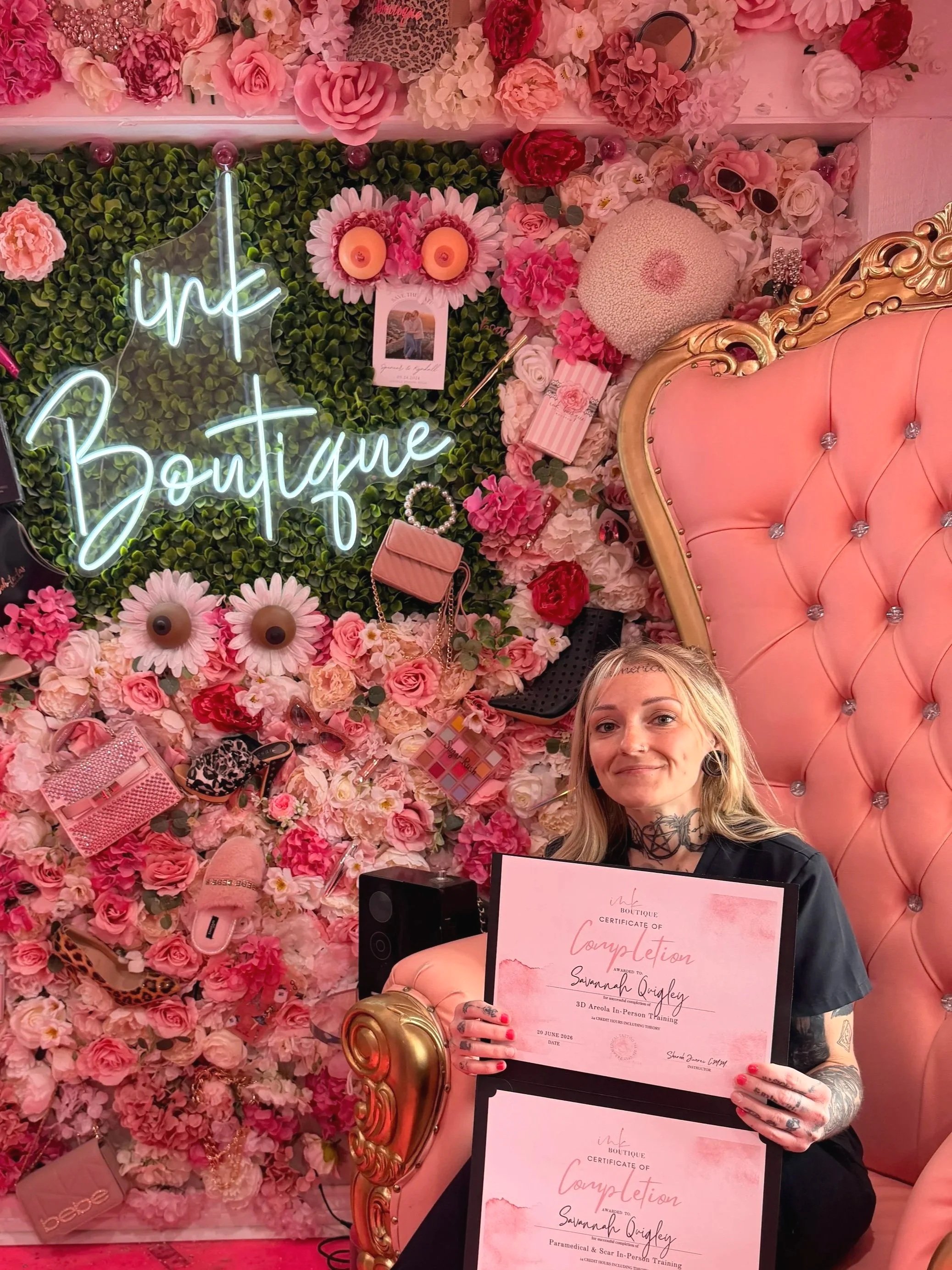 A woman sitting on a pink and gold ornate chair, holding a certificate of completion, in front of a wall decorated with pink and red flowers, a neon sign that says 'ink Boutique,' and various accessories like sunglasses and handbags.