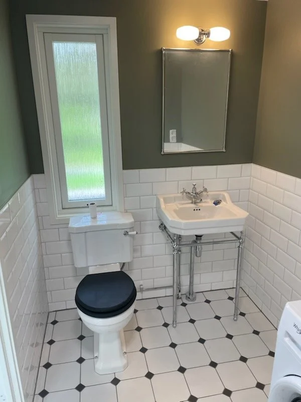 Liten badrum med vit toalett med svart sittlock, litet handfat med kromstativ, spegel och vägglampor, fönster med frostad glas, väggar halvt klädda med vita kakel och målade gröna, golv med svart och vit rutmönstrad plastmatta.