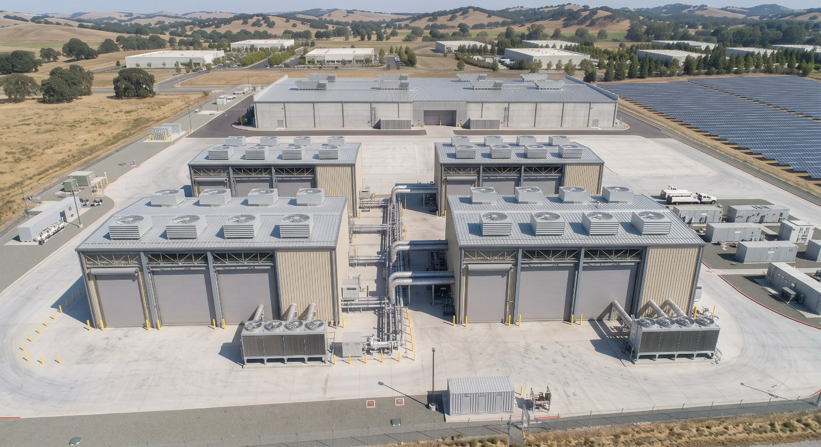 Large industrial facility with multiple buildings, ventilation units, and an array of solar panels on the adjacent land, surrounded by rural landscape.