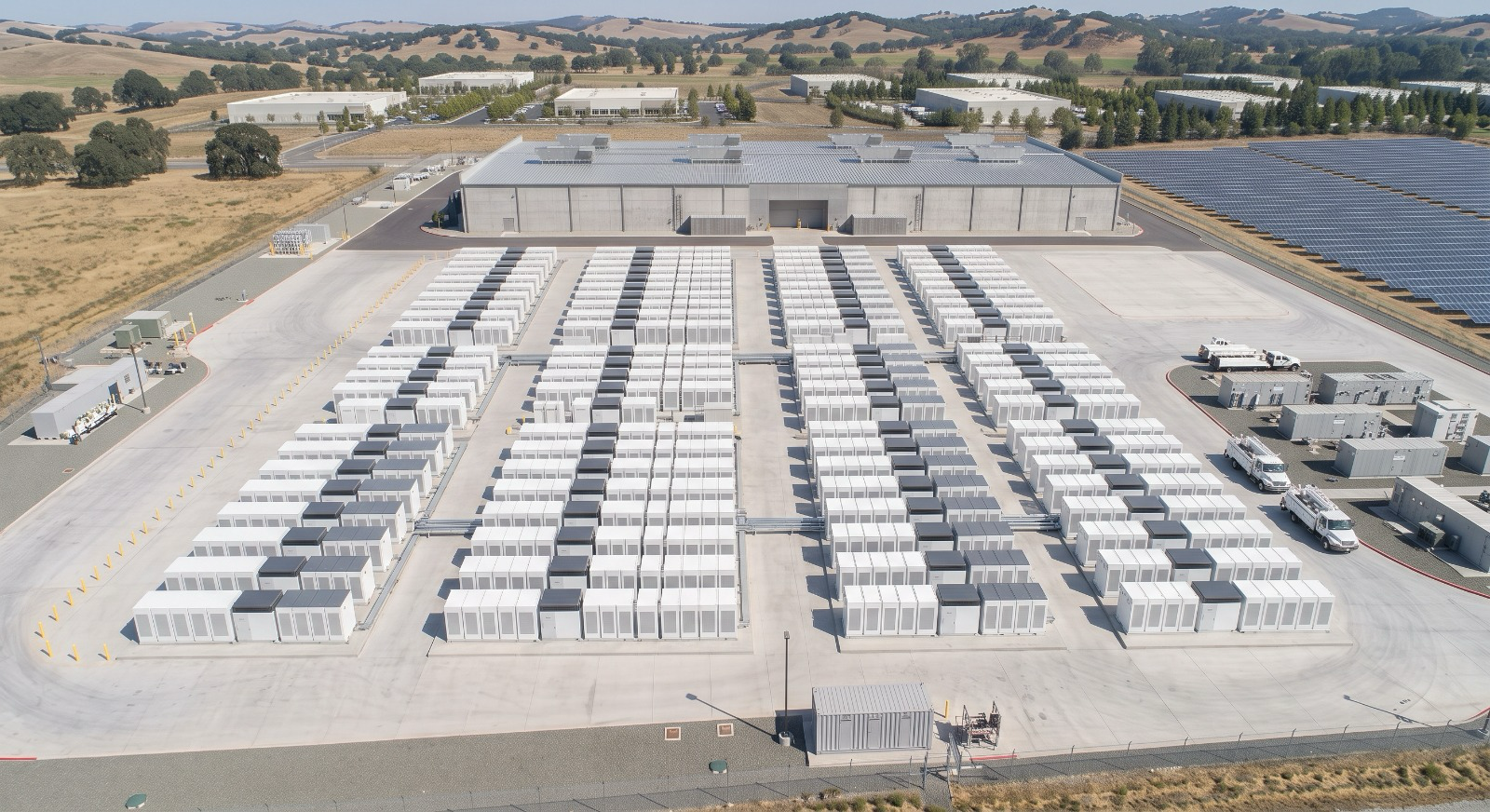 Aerial view of a data center with rows of cloud-like server units, solar panels on the right, and large industrial buildings surrounded by open land and hills in the background.