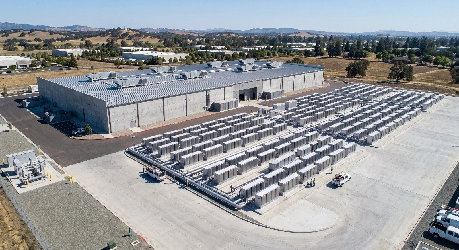 Aerial view of a large industrial building with a metal roof and numerous HVAC units on top, surrounded by a concrete parking lot and landscape with trees and rolling hills in the background.