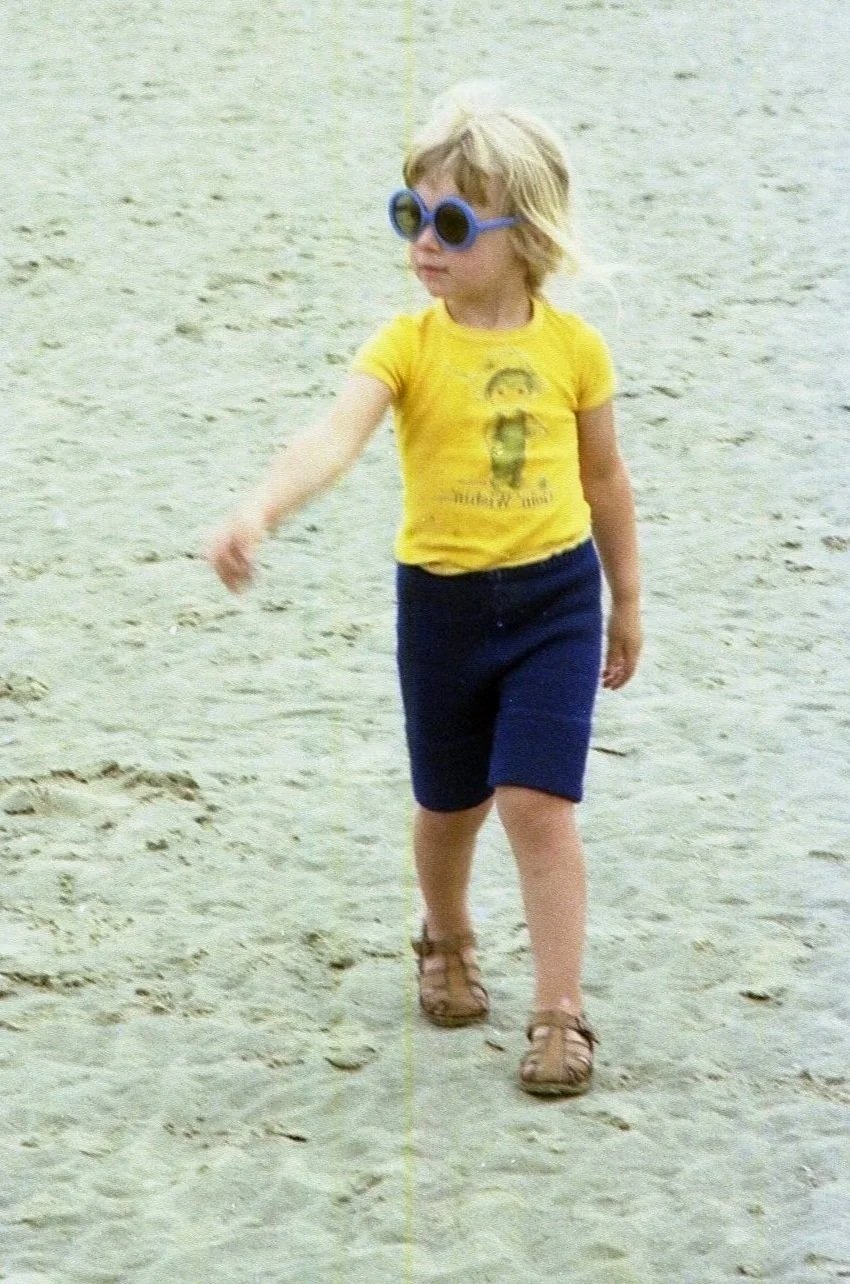 Kind met blond haar, grote blauwe zonnebril, gele T-shirt en blauwe shorts wandelt op zandstrand.
