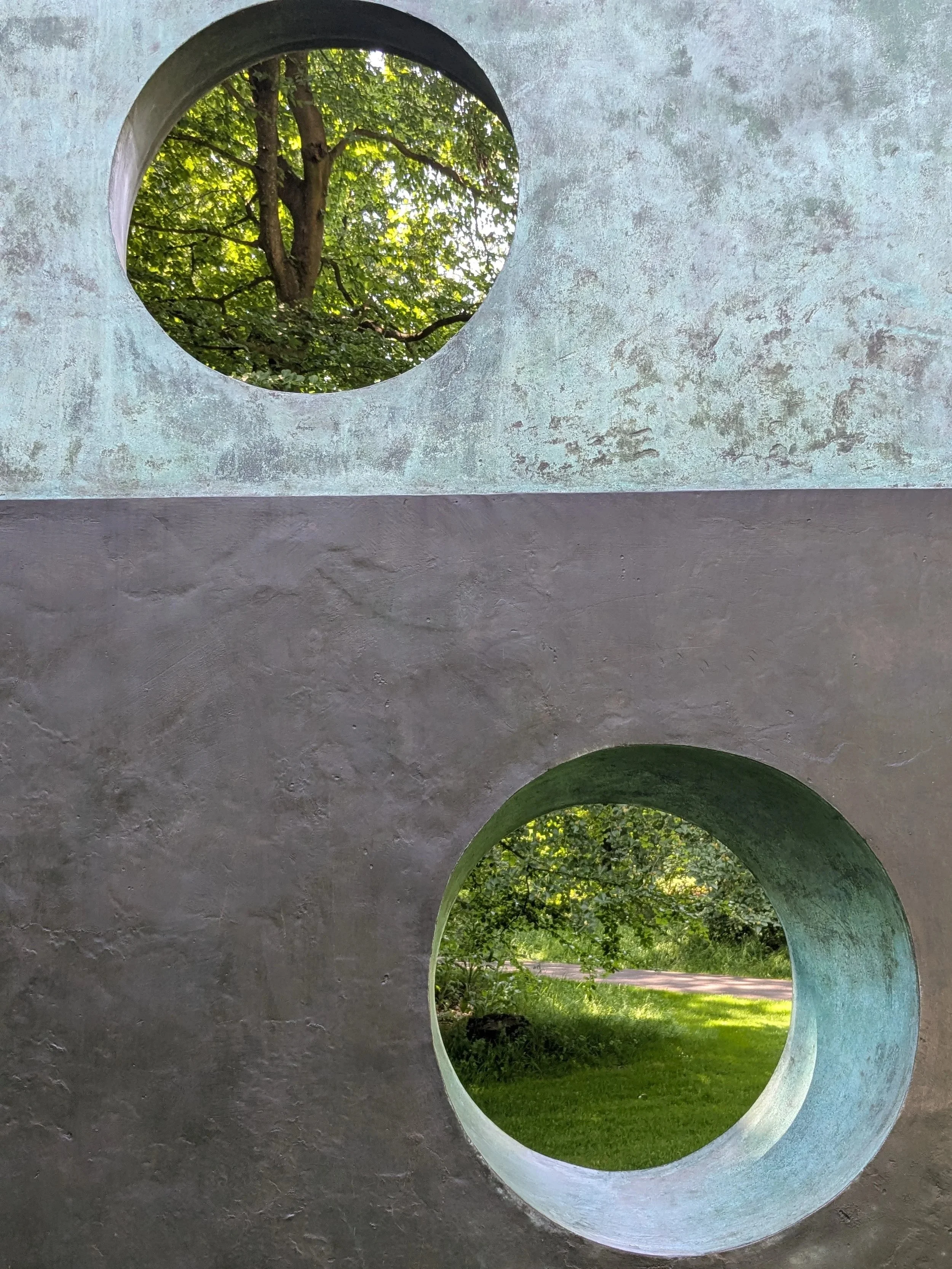 Een betonnen structuur met ronde openingen die uitzicht bieden op groene bomen en een grasveld.
