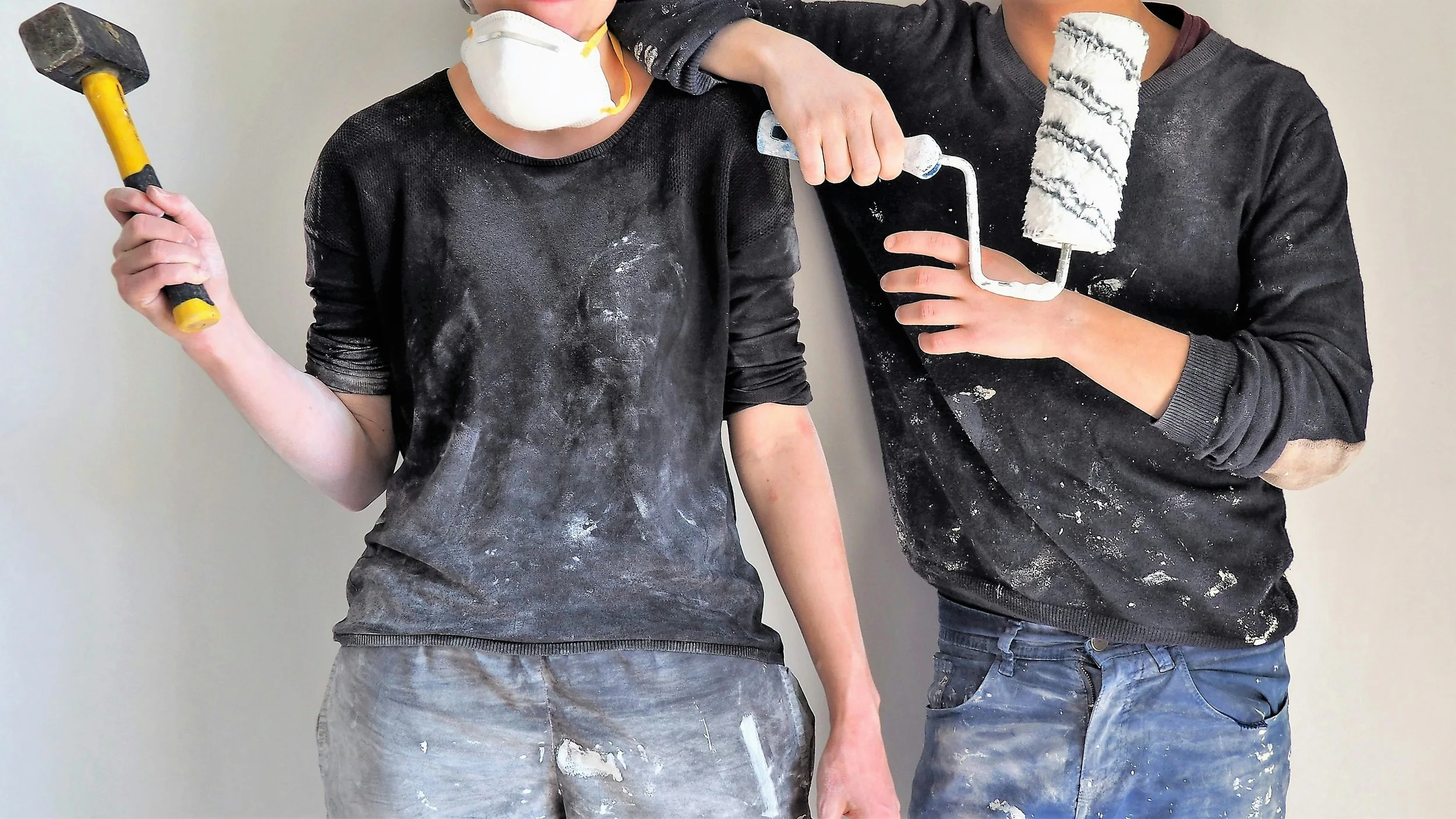 Two people wearing black shirts with paint stains, holding and using paint rollers, with white paint on their clothing and hands.