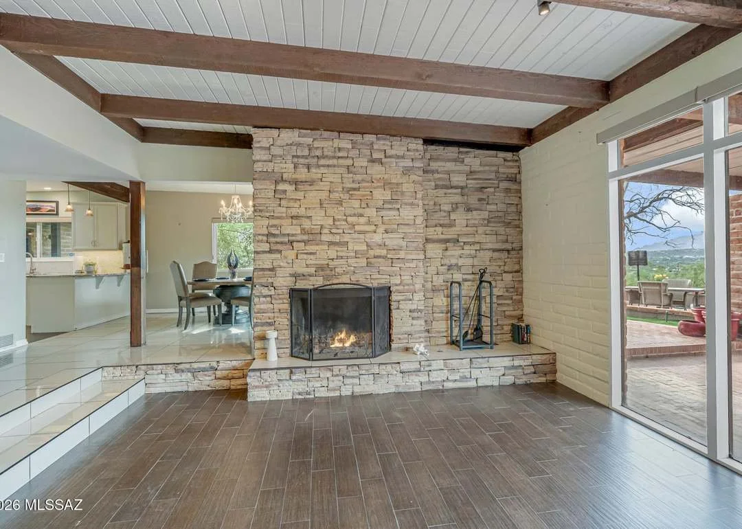 Living room with a stone fireplace, wooden beams on the ceiling, a view of the dining area and kitchen, and a sliding glass door leading to an outdoor patio with seating and a scenic view.