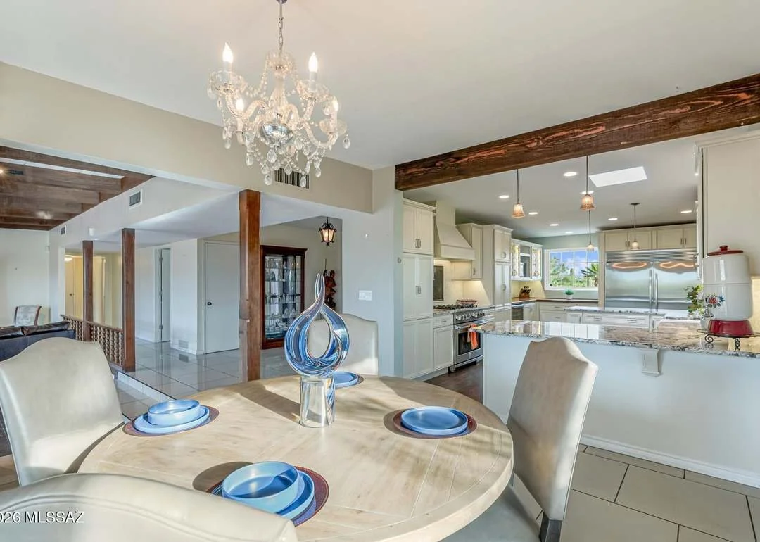 Dining area with a round table set with blue dishes, a modern glass sculpture centerpiece, cream-colored chairs, and a chandelier hanging above. The background features a spacious kitchen with white cabinets, granite countertops, and stainless steel appliances, illuminated by natural light from large windows.