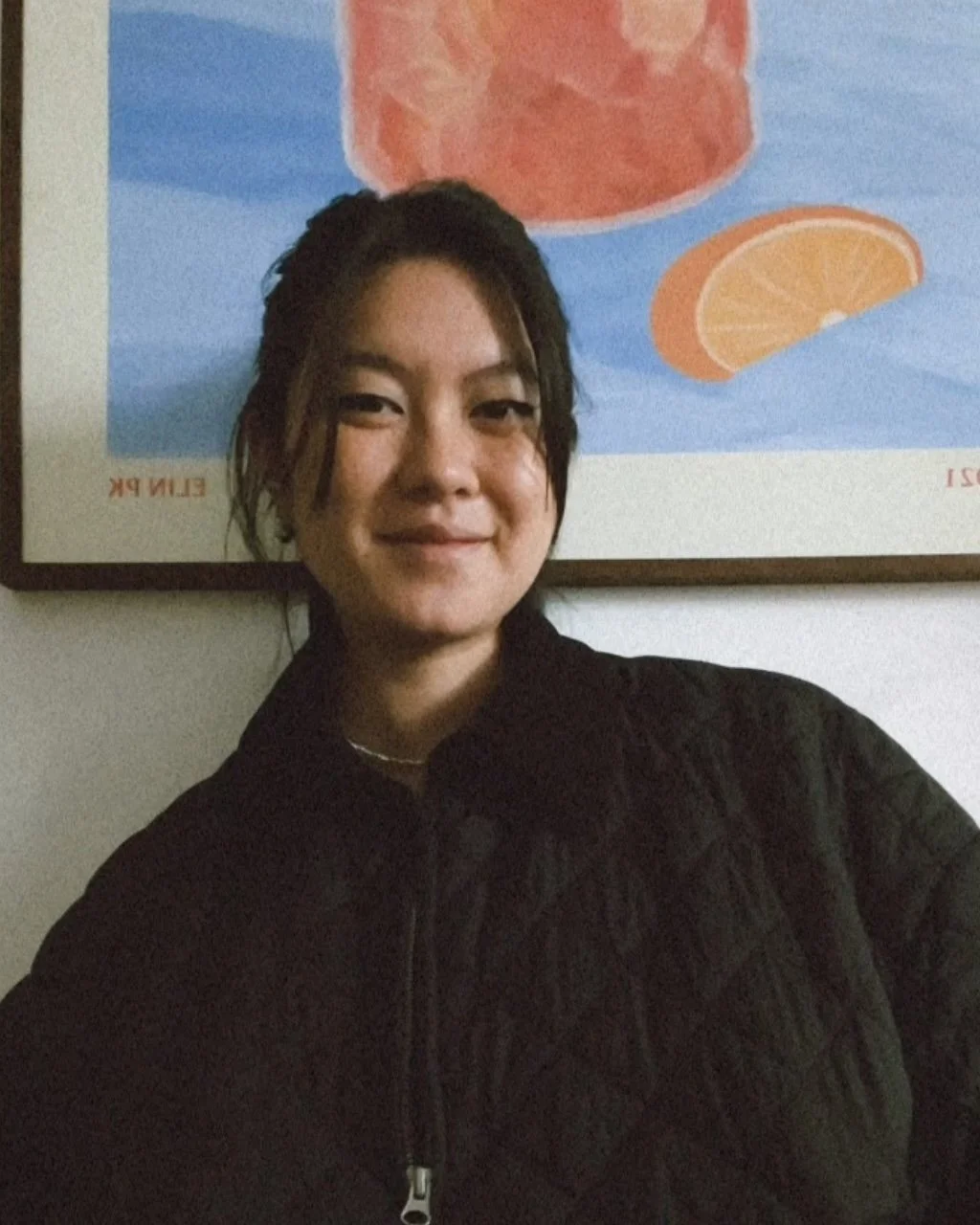 A woman with shoulder-length dark hair smiling, wearing a black quilted jacket, standing in front of a colorful poster of orange slices and a large glass of orange juice.