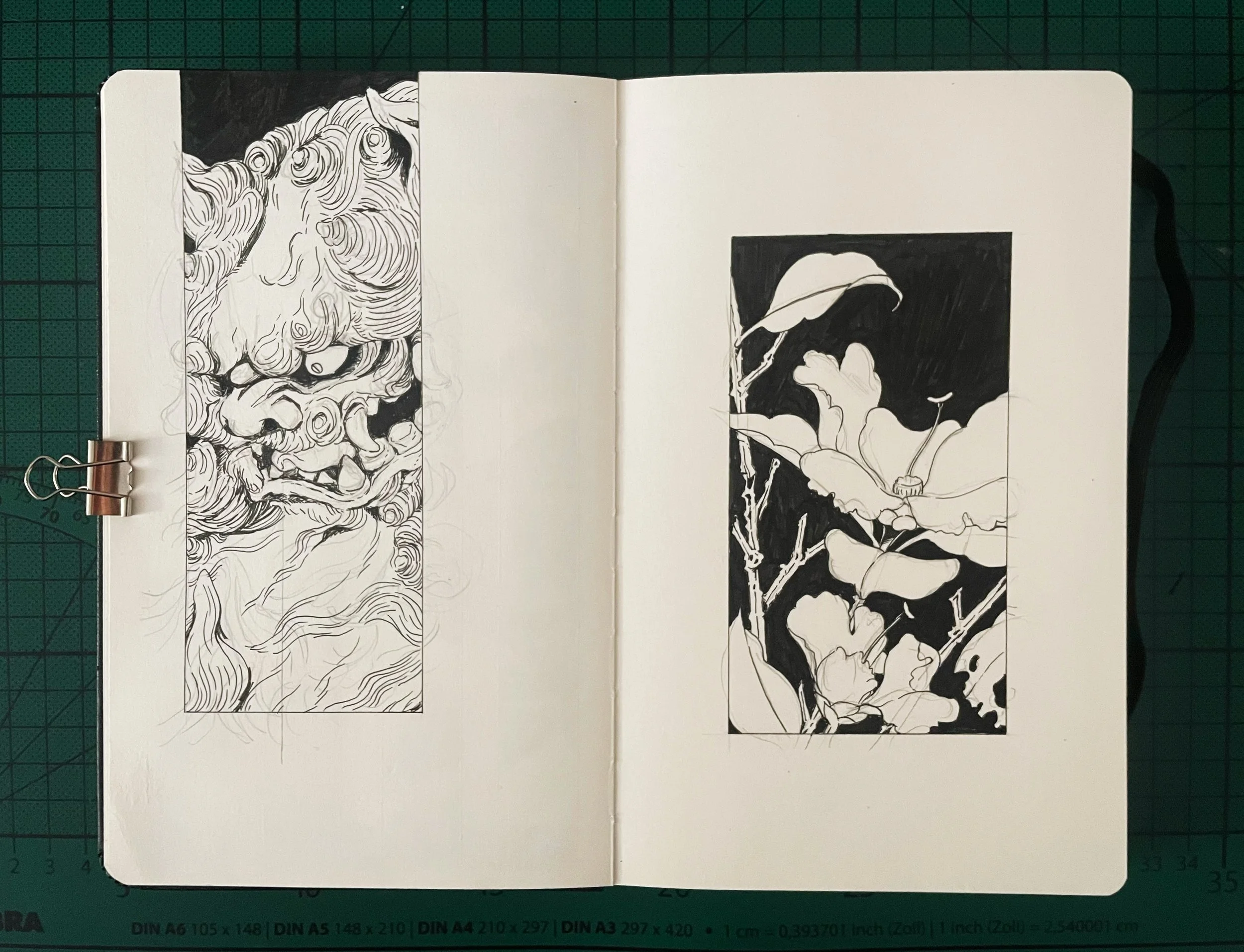Open sketchbook with black and white ink drawings of a lion's face on the left page and a plant with large leaves and flowers on a dark background on the right page.