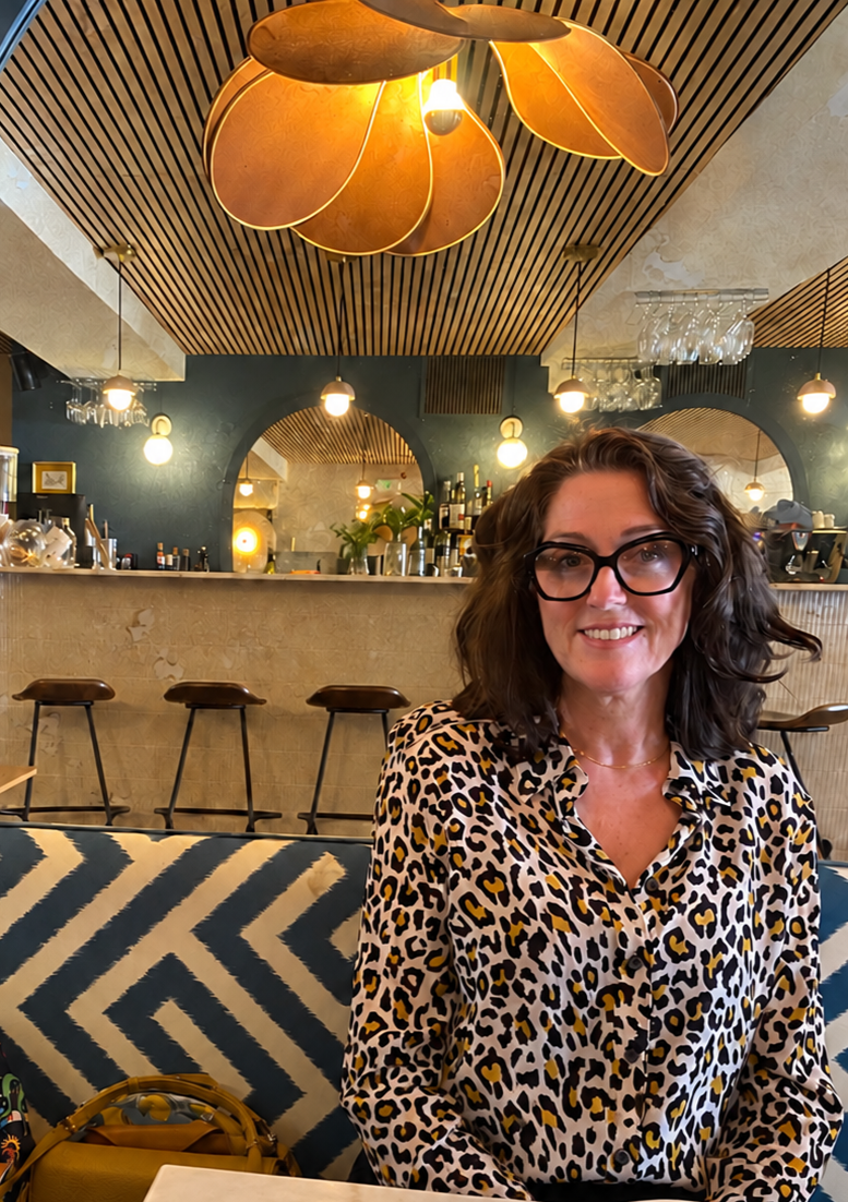 Une femme souriante portant des lunettes à motifs léopard, assise dans un café avec un décor moderne et chaleureux, un plafond en bois, un bar en arrière-plan, et un éclairage tamisé.
