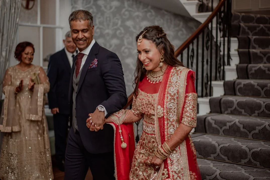 Claire Field Bridal Hair and Make up. A couple dressed in traditional Indian wedding attire, walking hand-in-hand down a staircase with guests clapping in the background.