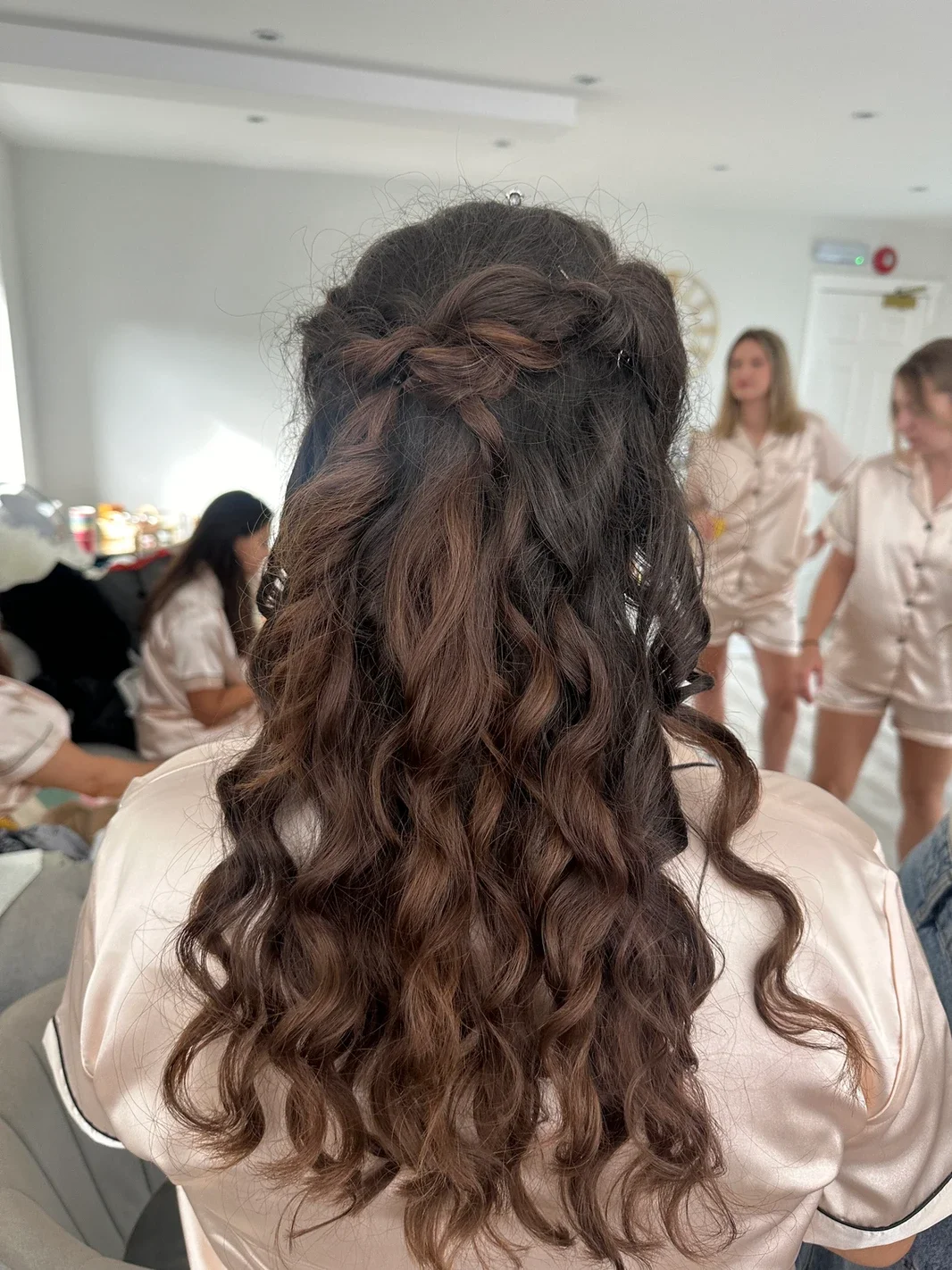 Claire Field Bridal Hair and Make up. Back of a woman with long, curly brown hair styled with a braid on top, wearing a satin pajama set, in a room with other women in matching pajamas, some sitting and some standing.