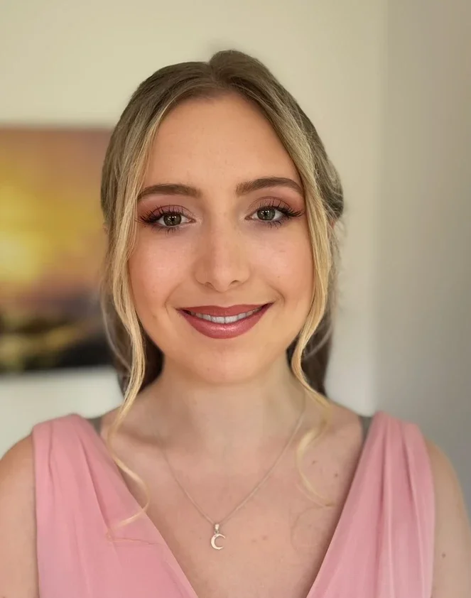 Claire Field Bridal Hair and Make up. A young woman with blonde hair styled in loose curls, wearing a pink sleeveless top and a moon necklace. She is smiling and standing indoors with a blurred background.
