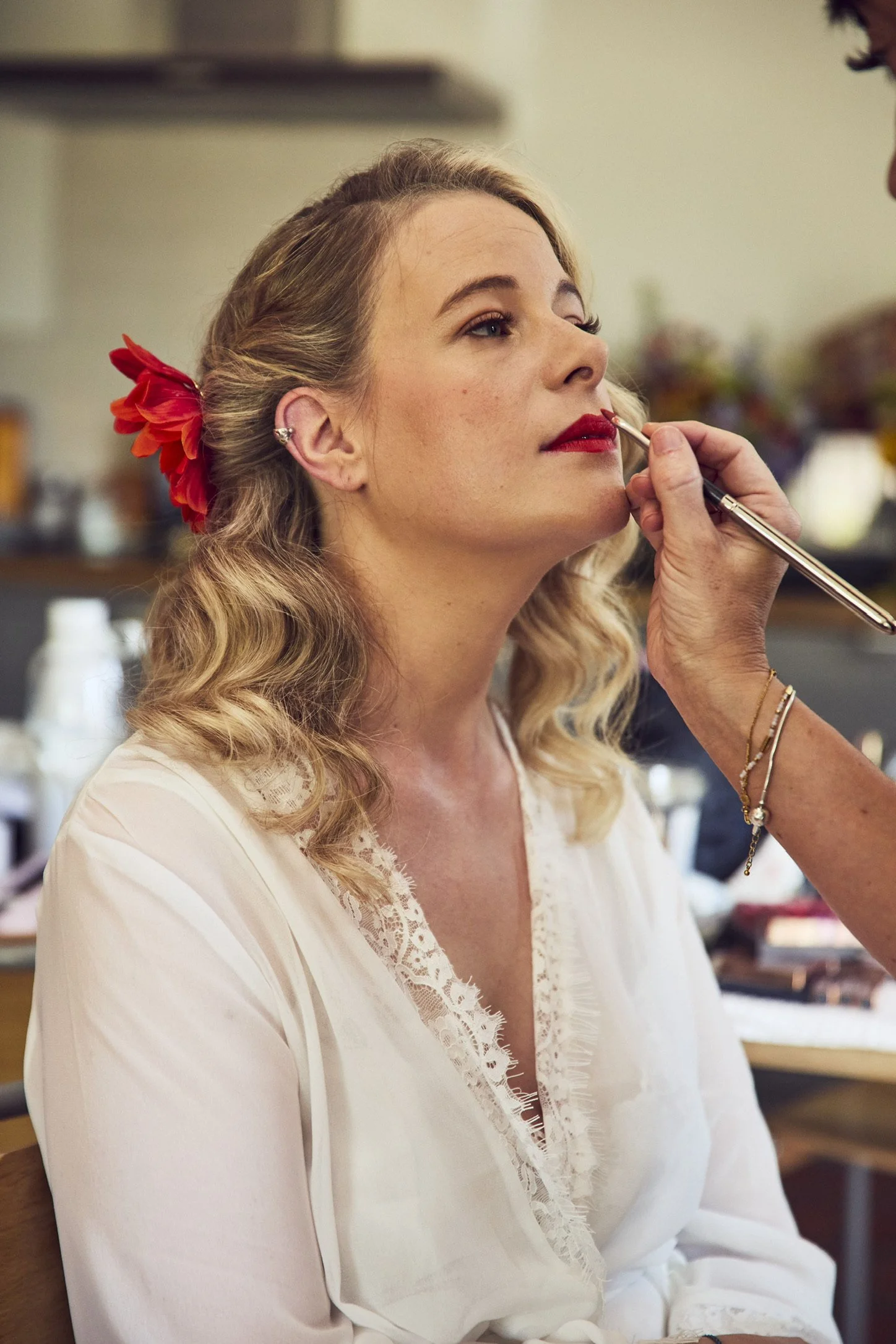 Claire Field Bridal Hair and Make up. A woman with blonde hair, a large red flower in her hair, wearing a white blouse with lace detailing, has lipstick applied by a makeup artist, in a cozy, decorated room.
