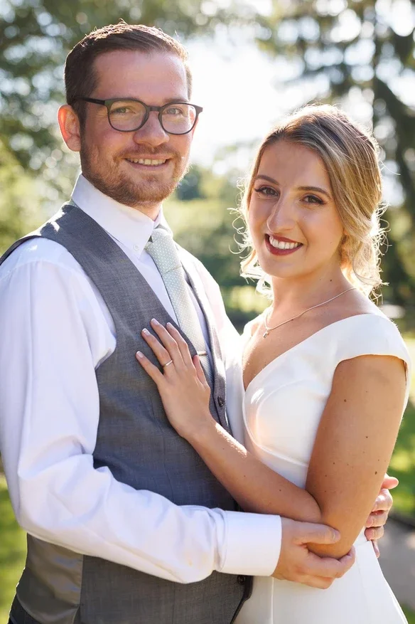 Claire Field Bridal Hair and Make up. A smiling couple outdoors with trees in the background, dressed in formal attire; the woman is wearing a white dress and the man is in a white shirt with a gray vest and tie, embracing each other.