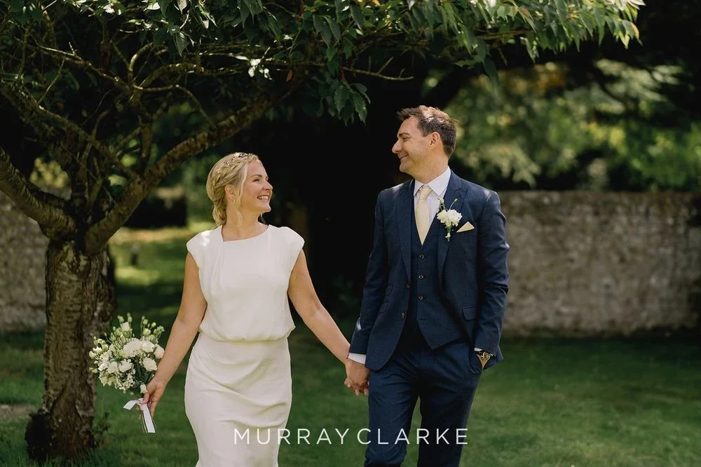 Claire Field Bridal Hair and Make up. A bride and groom walking hand in hand outdoors, smiling at each other during a wedding. The bride is holding a bouquet of white flowers, and they are under a tree with a stone wall in the background.