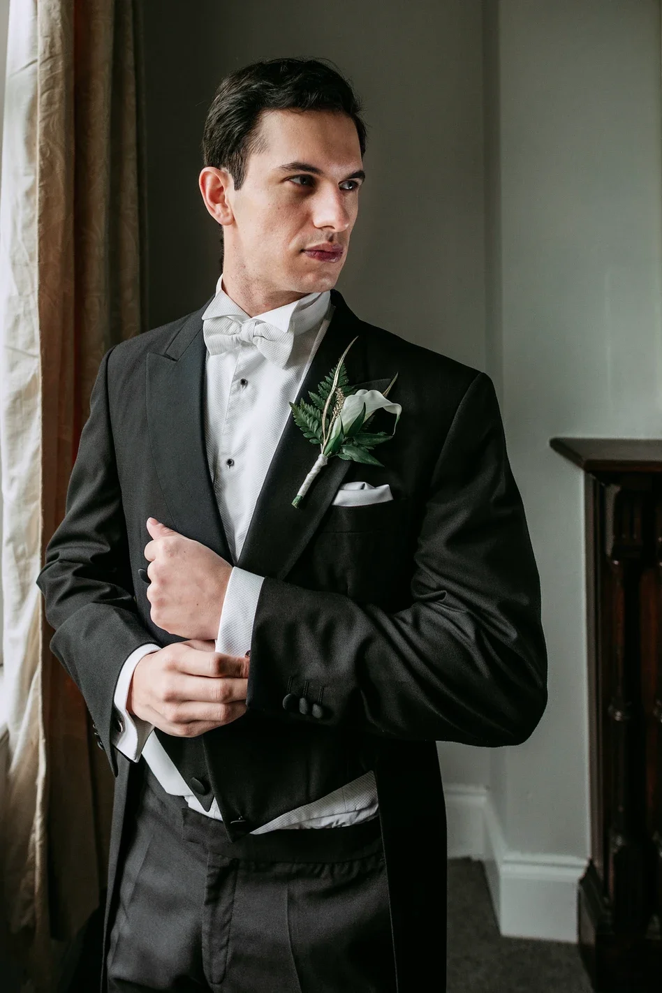 Claire Field Bridal Hair and Make up. A man dressed in a tuxedo with a white bow tie and boutonniere standing indoors near a window with curtains.