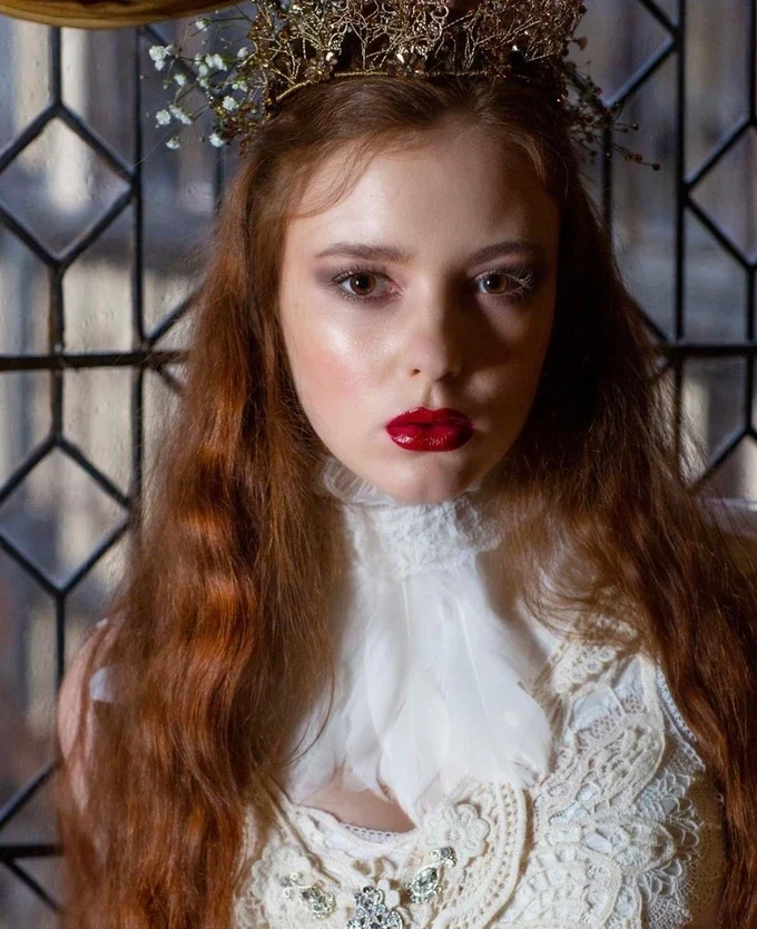 Claire Field Bridal Hair and Make up. Young woman with long red hair wearing a white lace blouse and a decorative crown or tiara, standing in front of a black metal fence.