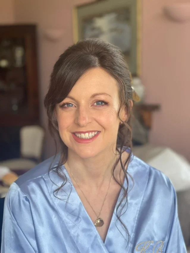 Claire Field Bridal Hair and Make up. A woman with light skin, blue eyes, and brown hair styled in an updo with loose curls, smiling. She is wearing a light blue satin robe with embroidery and a gold necklace. 