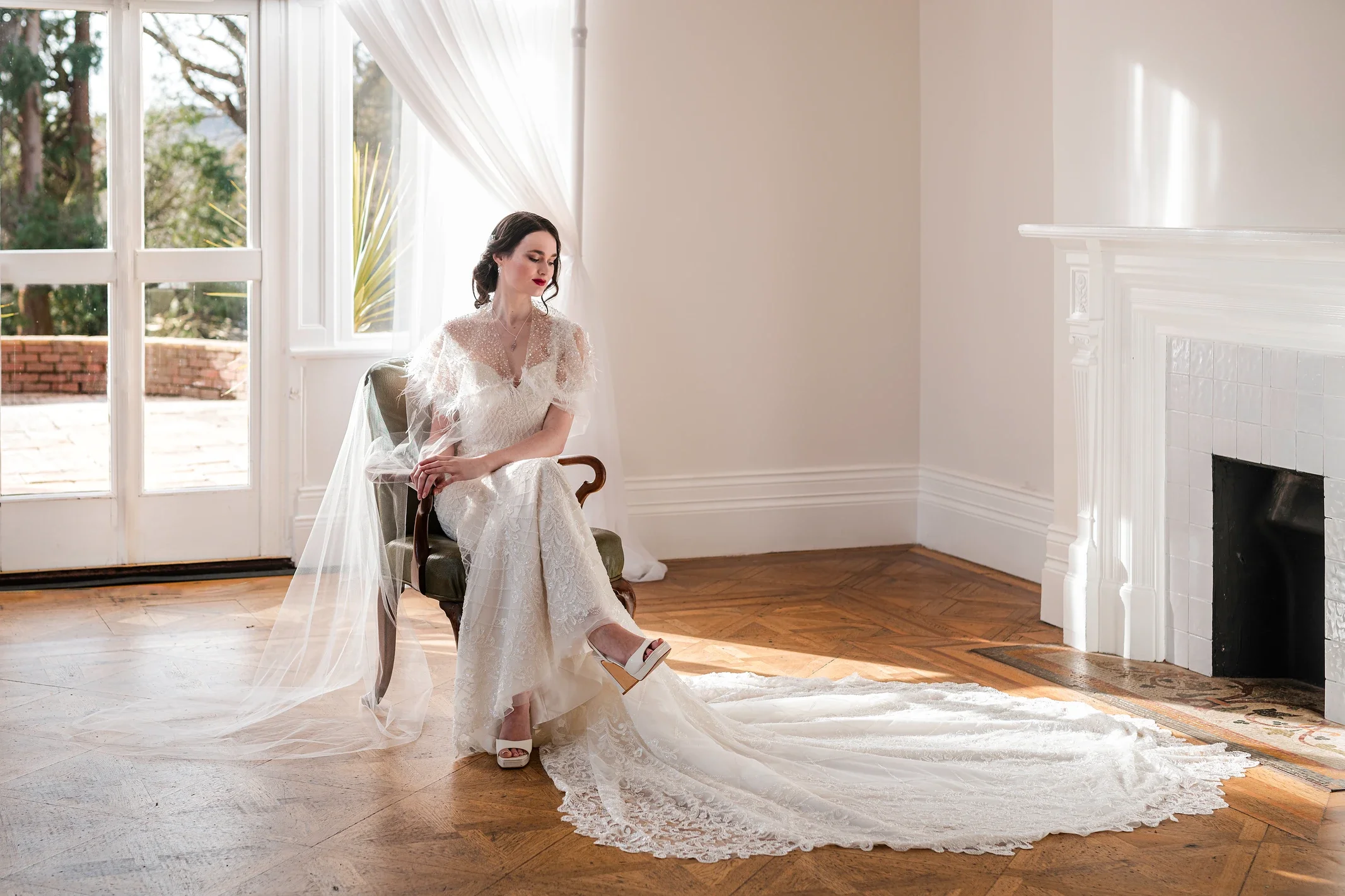 Claire Field Bridal Hair and Make up. A woman in a white lace wedding dress with a long train sits on a vintage armchair in a bright room with large windows, a white fireplace, and wood floors, looking downward with a serene expression.